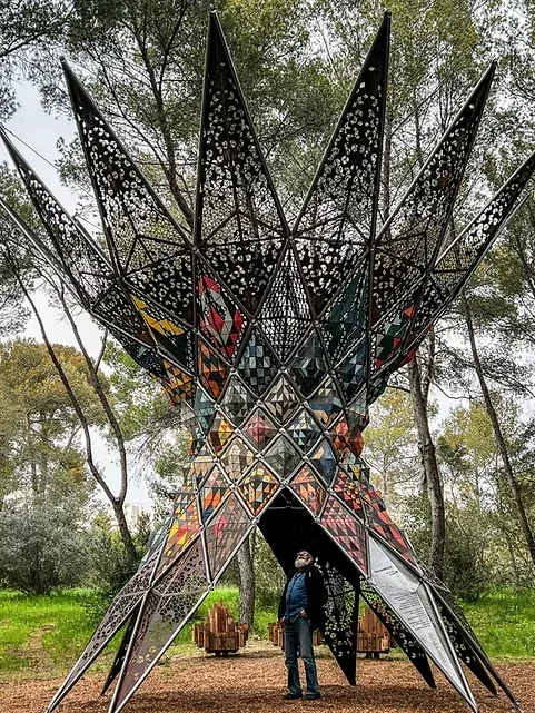 Sculpture resembling a large, star-shaped, spiked structure made of metal and stained glass panels, located outdoors in a wooded area with a person standing underneath.