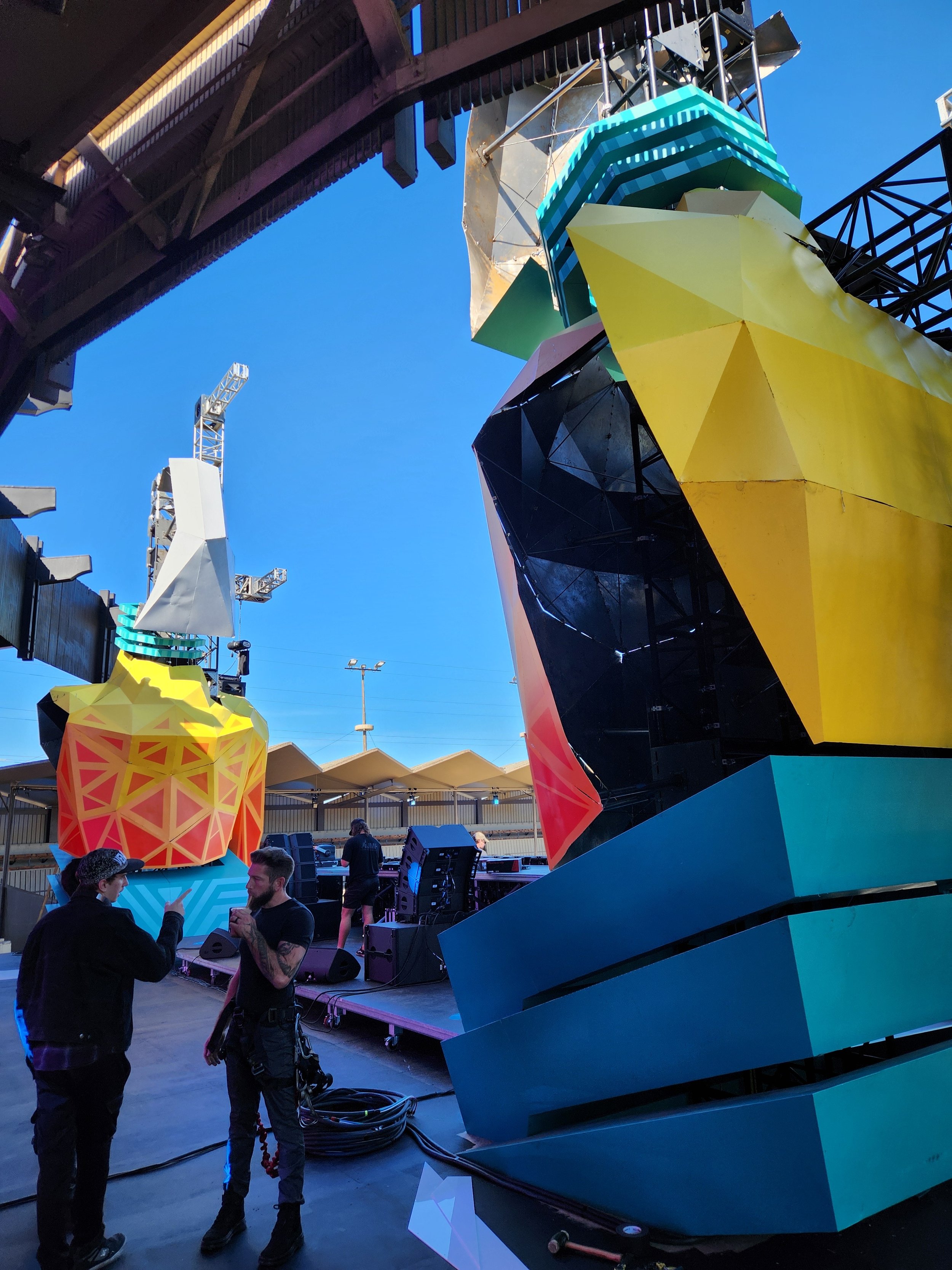 People standing and talking in front of an installation with vibrant, colorful geometric shapes and black scaffolding at an outdoor event under a clear blue sky.