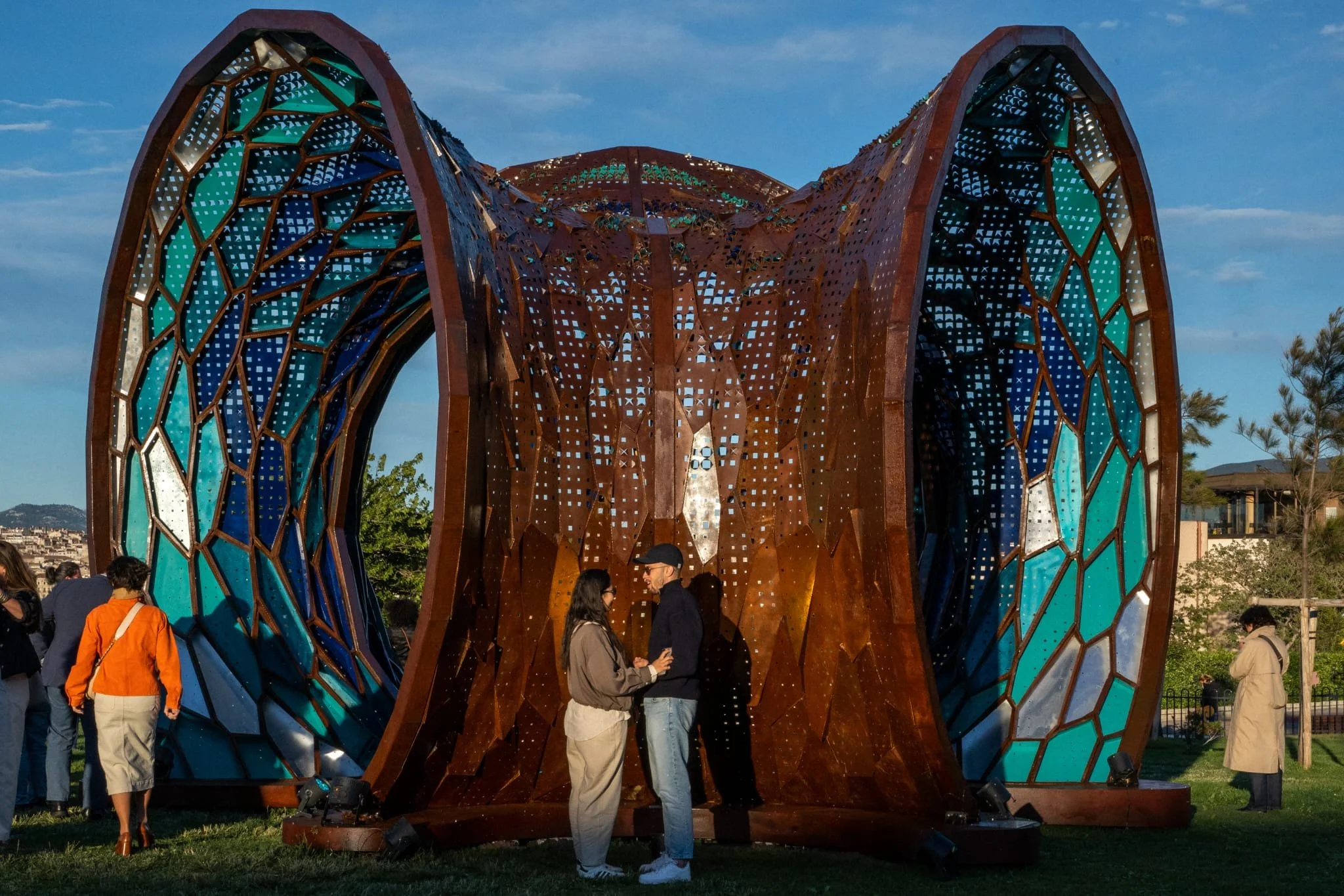 People gather around and under a large, artistic metal structure with a curved, open design, set outdoors during the day with a clear blue sky.