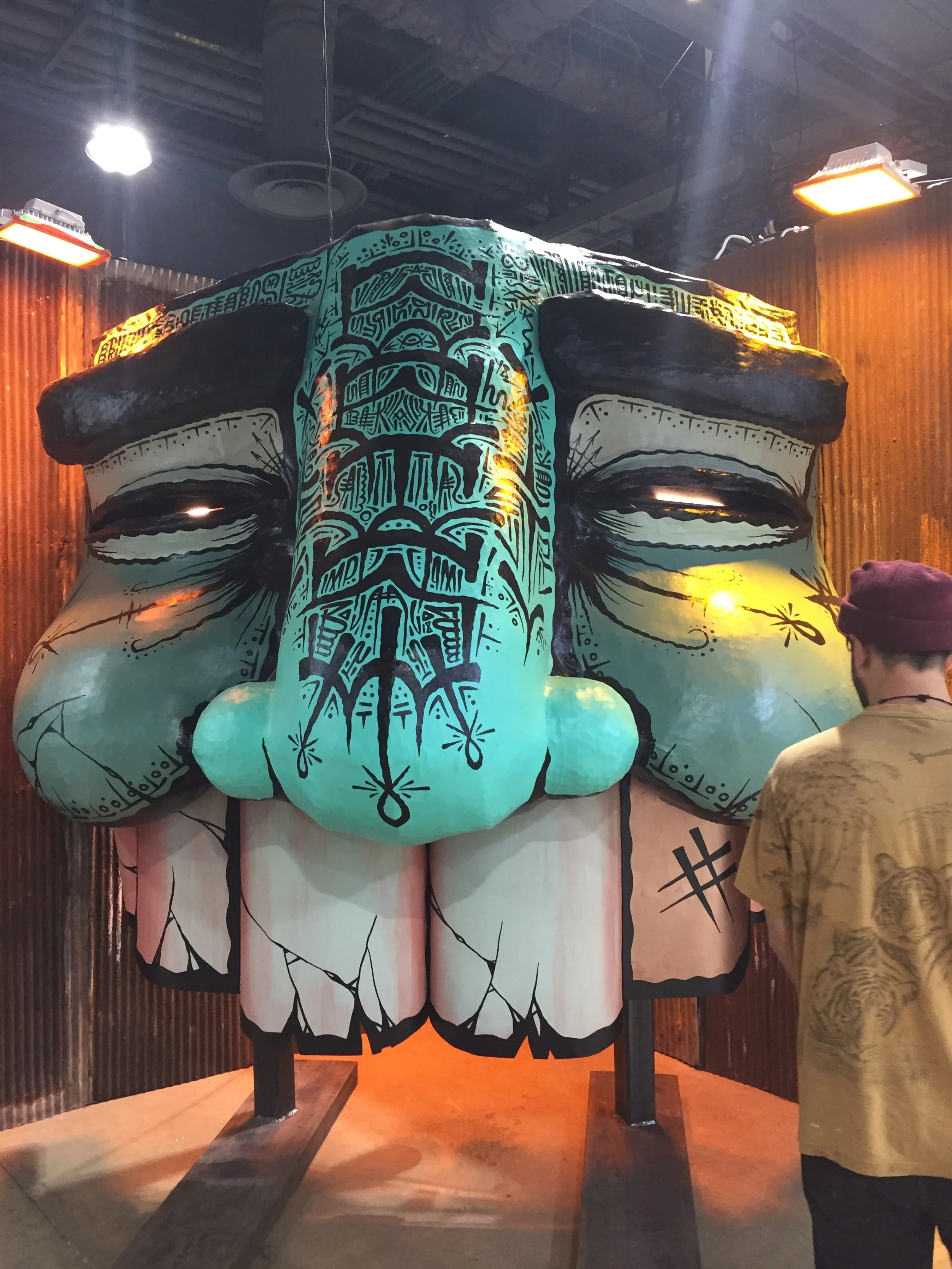 A large sculpture of a cartoonish face with a blue head, black eyebrows, and closed eyes, decorated with tribal-style patterns, is displayed indoors with wooden walls. A person in a beige shirt is standing nearby.