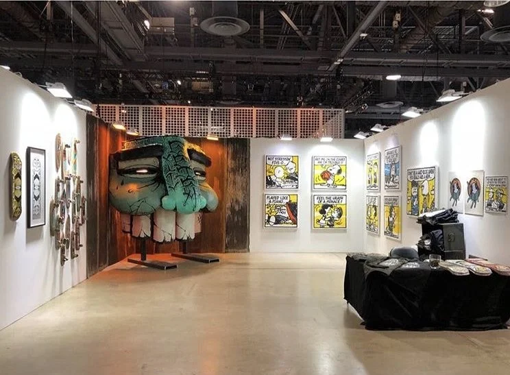 Art gallery with comic strip panels on the wall, a large colorful mask of a face with exaggerated features, and a table covered with various items.