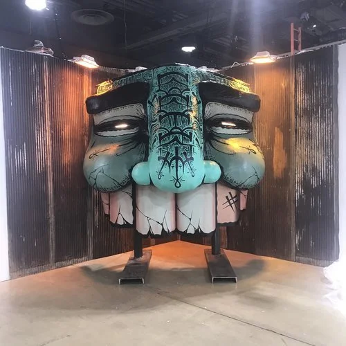 A large, colorful sculpture of a smiling cartoon face with exaggerated features, including a prominent nose, large eyelids, and a wide toothy grin, standing against a black corrugated metal wall.