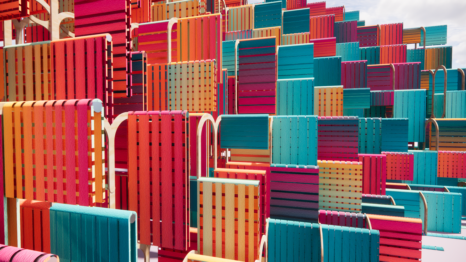A colorful outdoor installation composed of numerous stacked wooden chairs painted in bright shades of red, blue, orange, pink, and yellow, arranged in a layered, overlapping pattern against a partly cloudy sky.