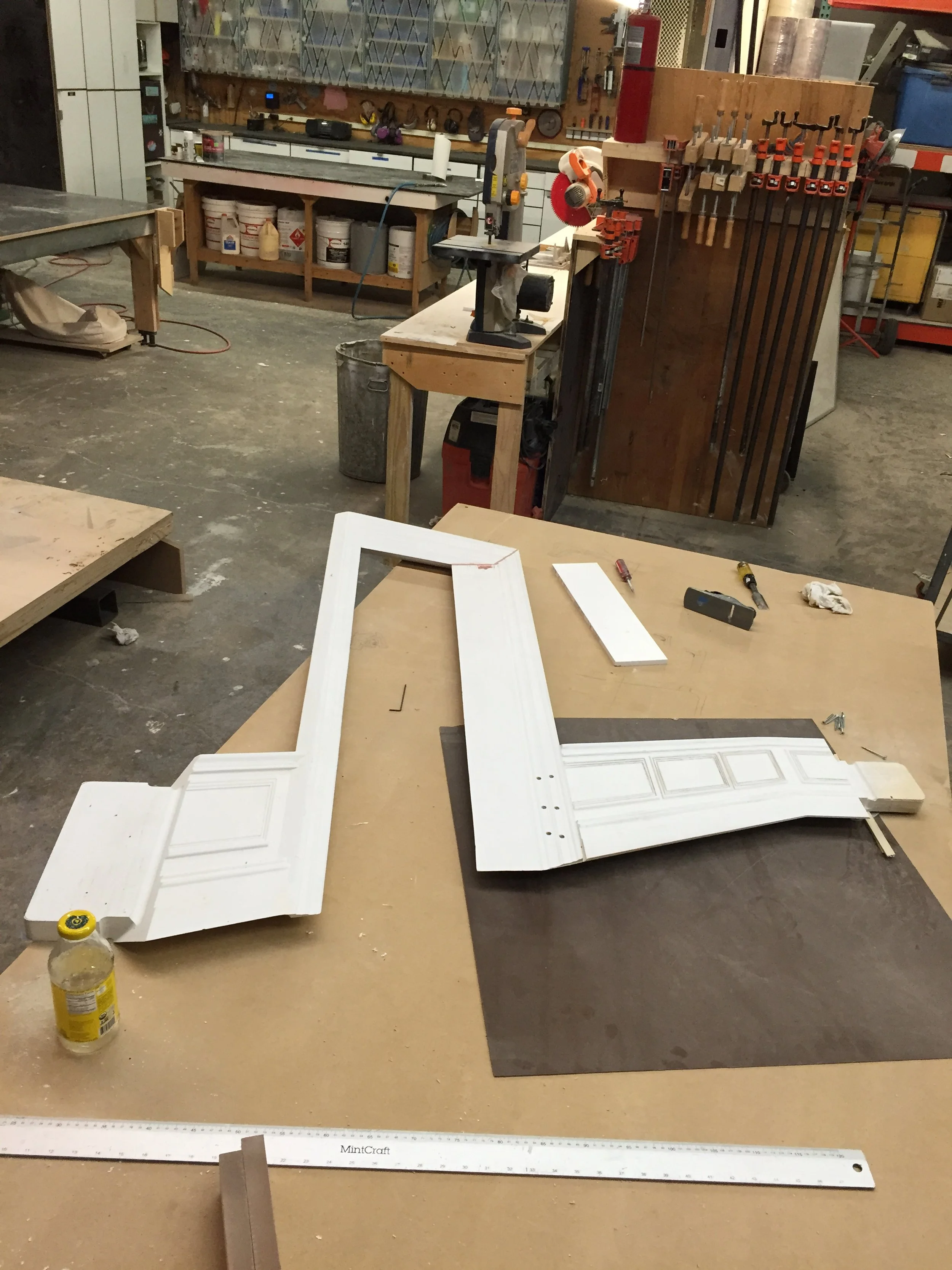 Workshop with tools and woodworking materials, including a white picture frame on a workbench, clamps, a measuring tape, and various power tools, with shelves and storage in the background.