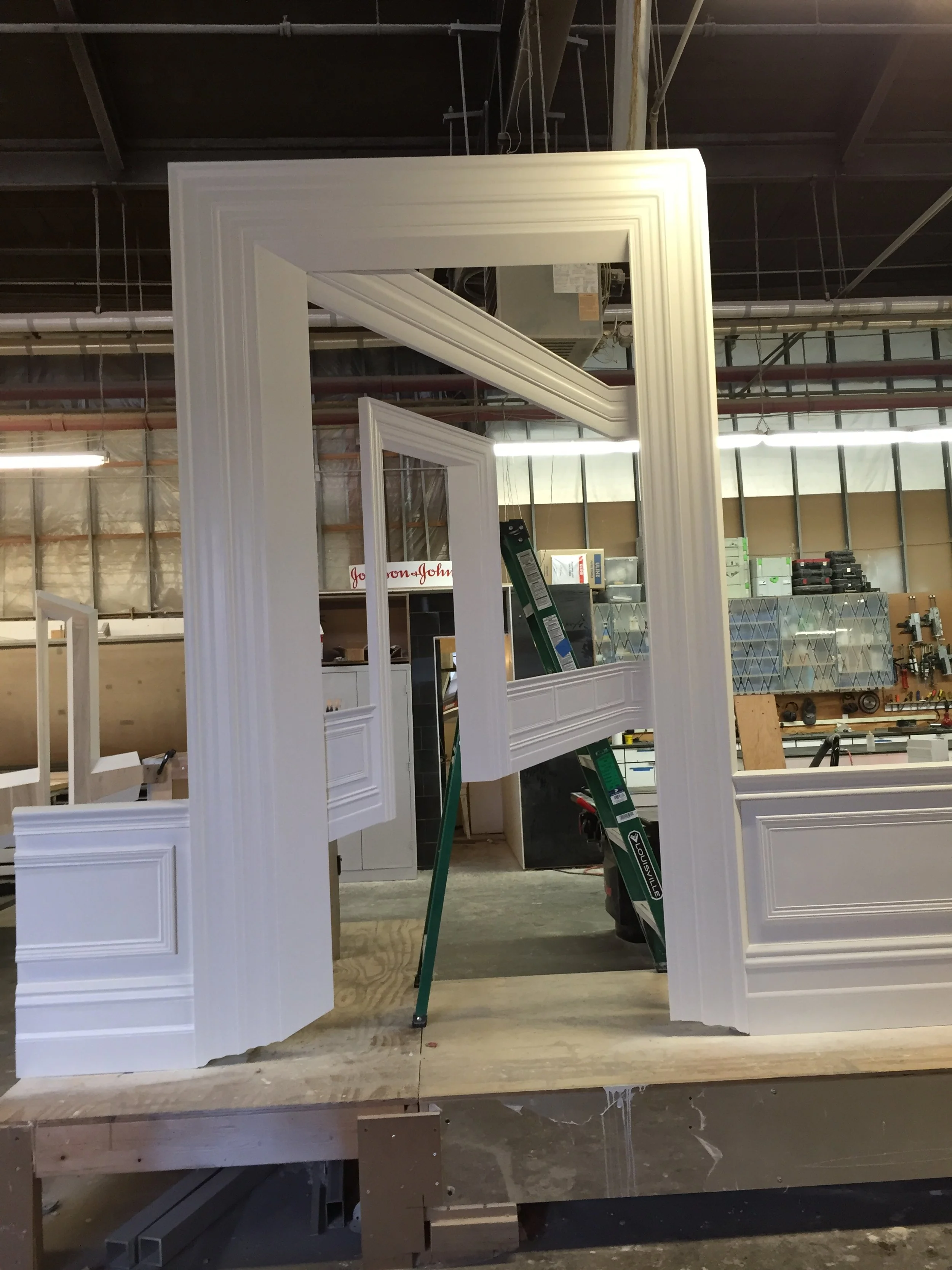 Warehouse setting with multiple white picture frames or moldings leaning against a workbench. In the background, there are tools, storage bins, and industrial pipes.