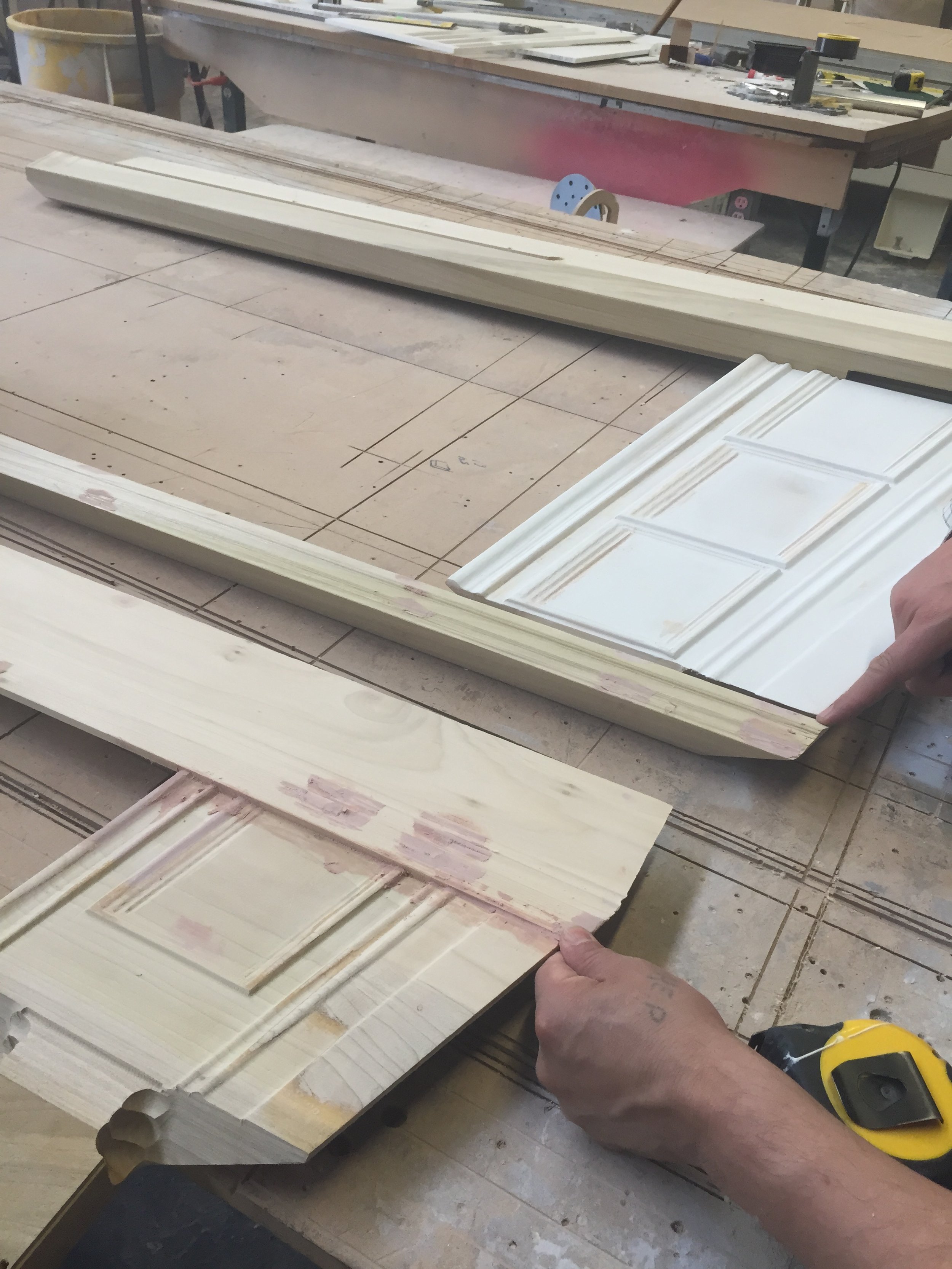 A person measuring decorative trim moldings with a yellow tape measure in a woodworking workshop.