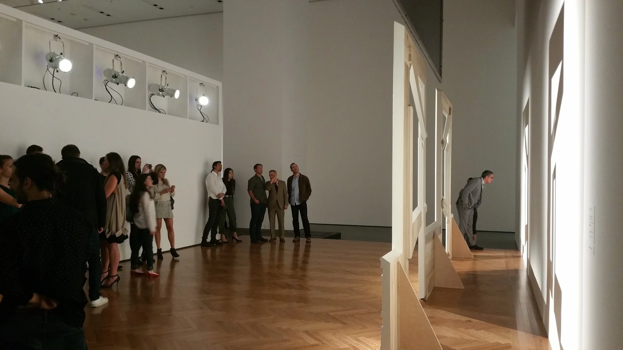 People gathered in an art gallery observing a display of lights and artwork on the wall.
