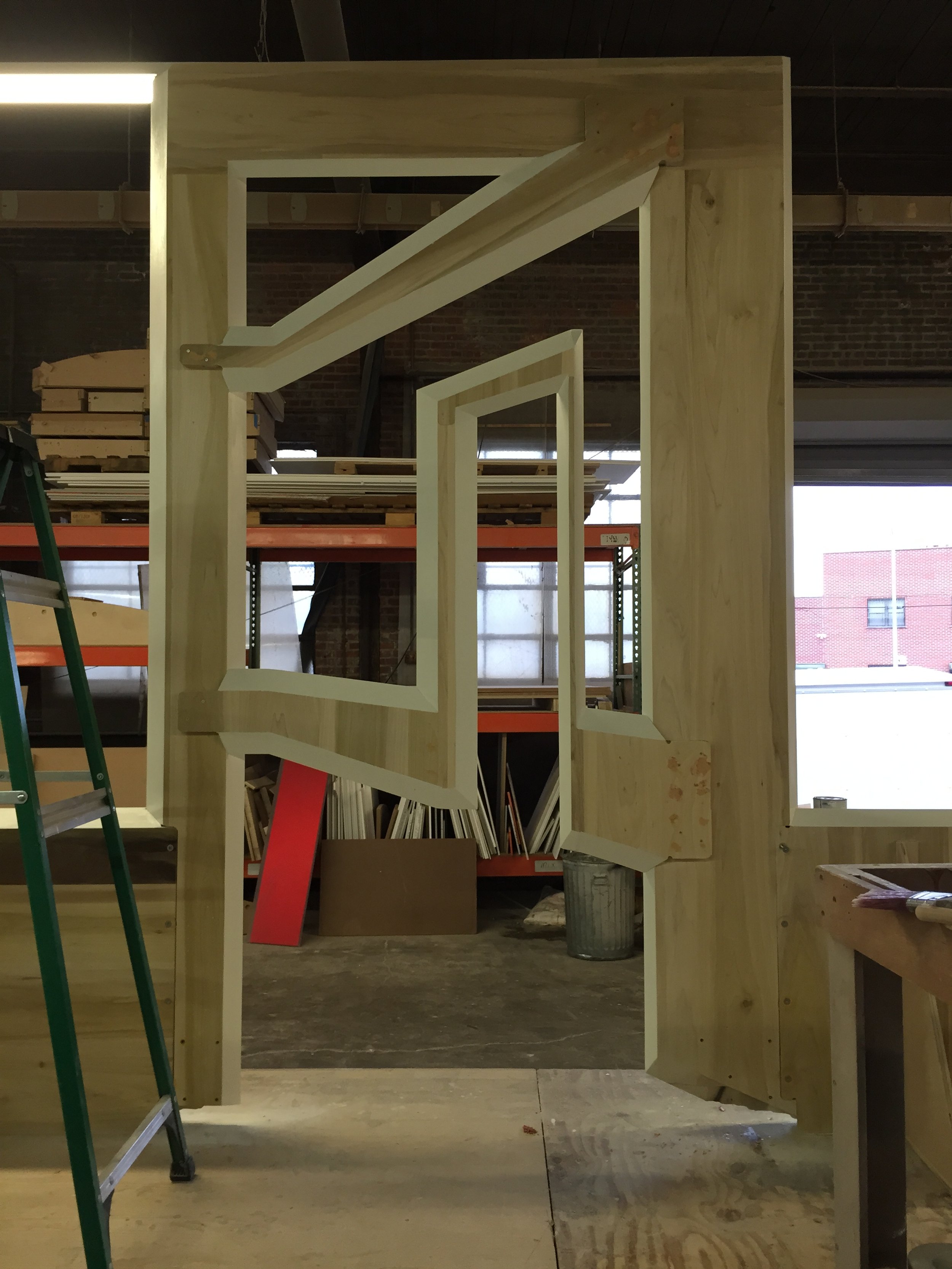 Construction site with wooden framing, mirror, ladder, and shelves in background, possibly in a workshop or building under construction.