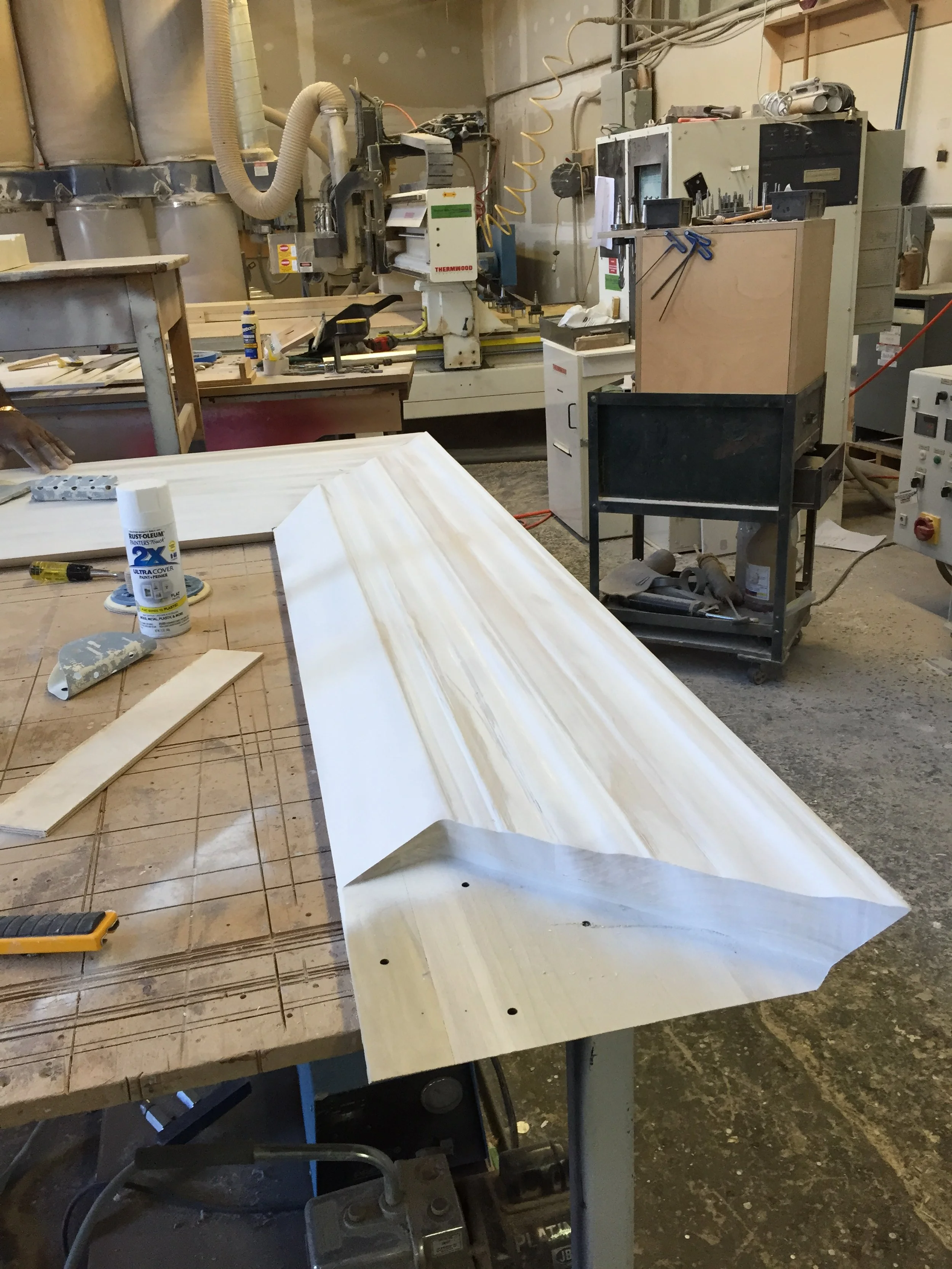 A woodworking workshop with a large piece of wood on a worktable, various tools, and equipment including a saw, clamps, and power cords. There is a machine labeled 'THERMWOOD' in the background, and a black chalkboard or sign leaning against a cart.