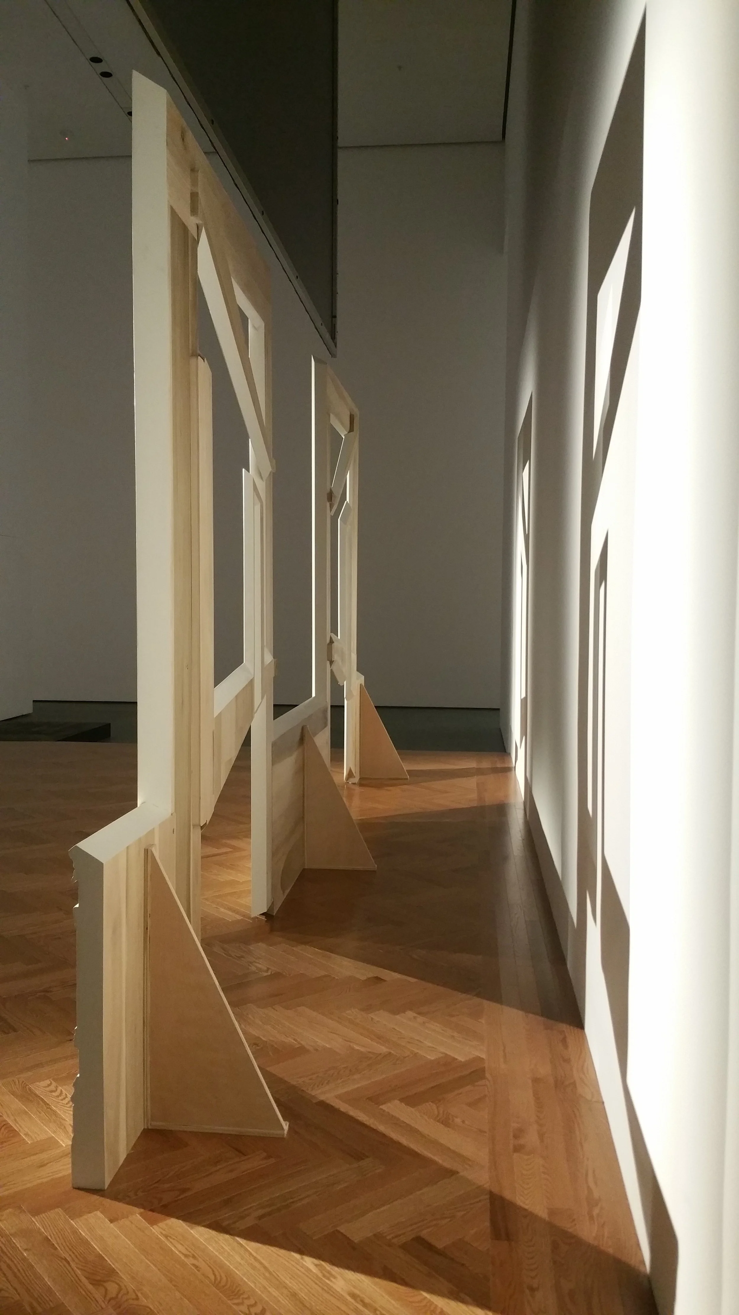 An art installation in a gallery with white and wooden panels that create shadow patterns on the wall. The panels are on easels and set on wooden flooring.