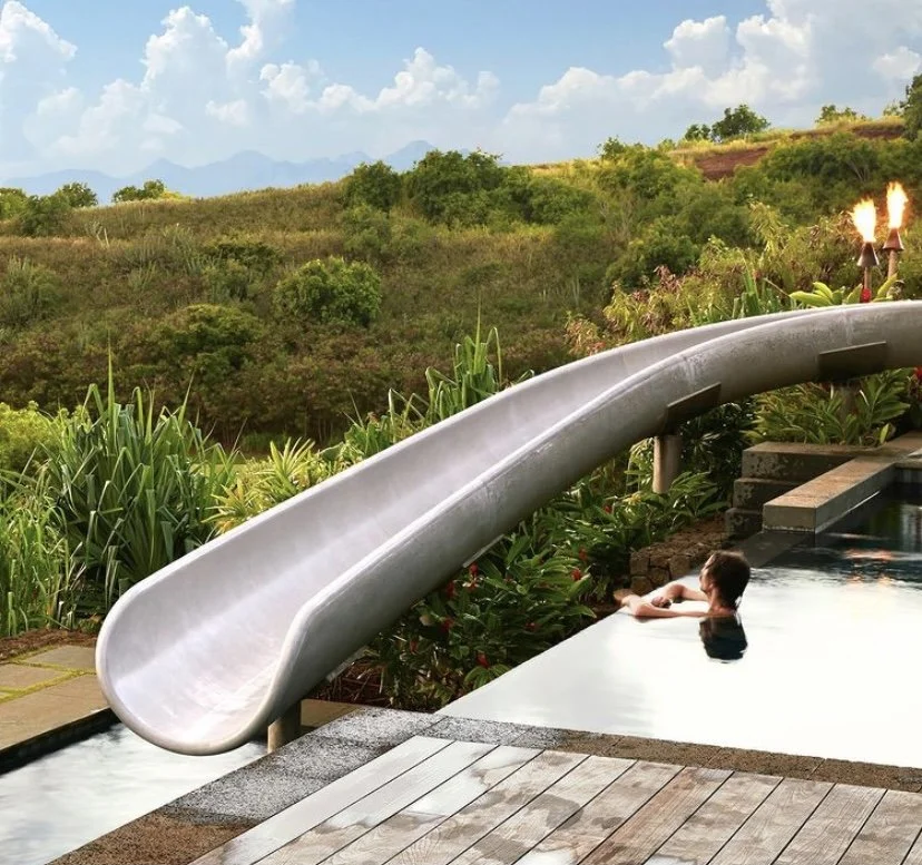Person relaxing in a pool with a water slide, surrounded by lush green plants, with a scenic mountain view in the background and flames in the distance.