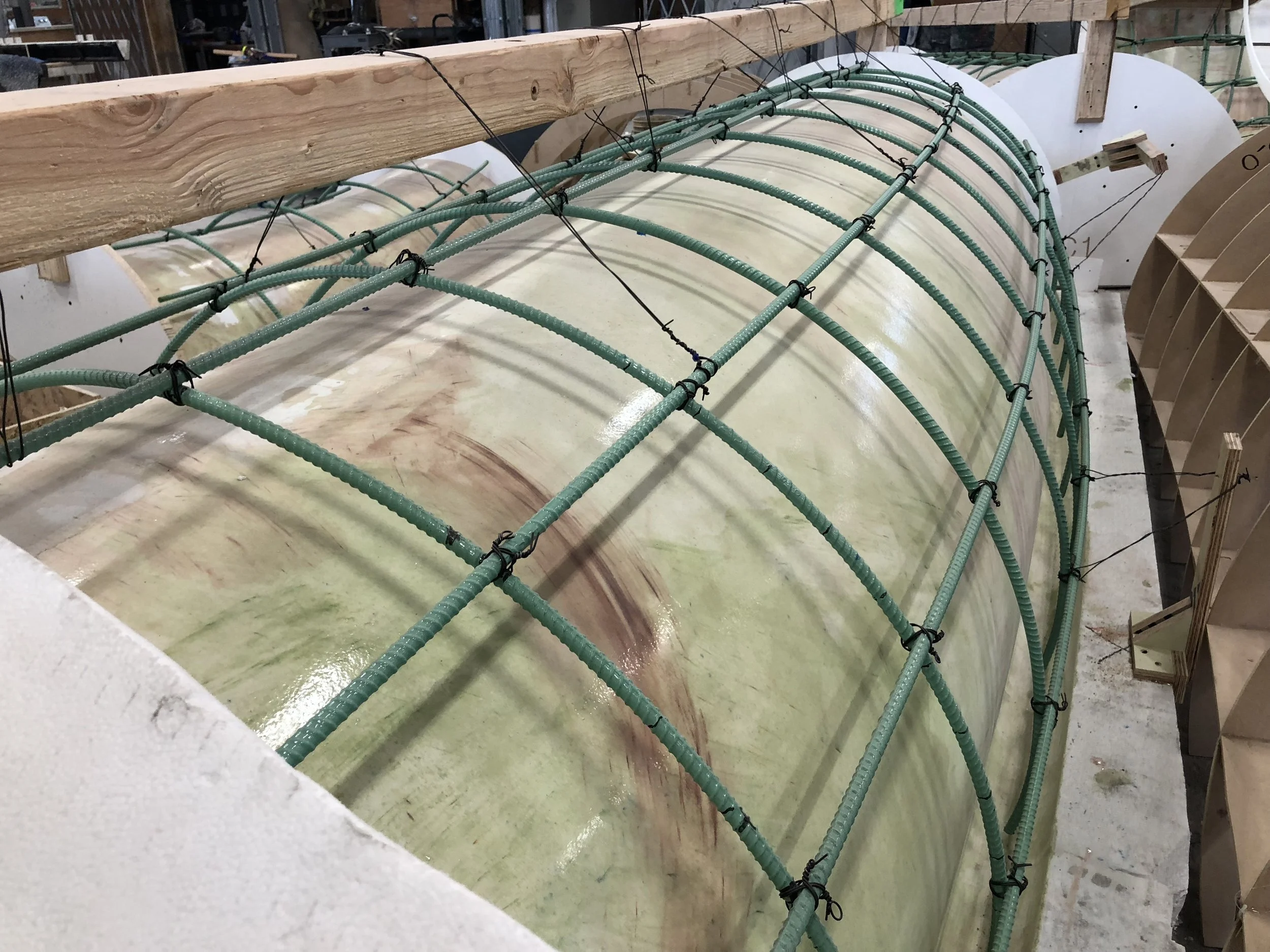 Close-up view of a fiberglass or resin curved surface with secured green hoses and black zip ties, supported by wooden beams in an industrial or workshop setting.