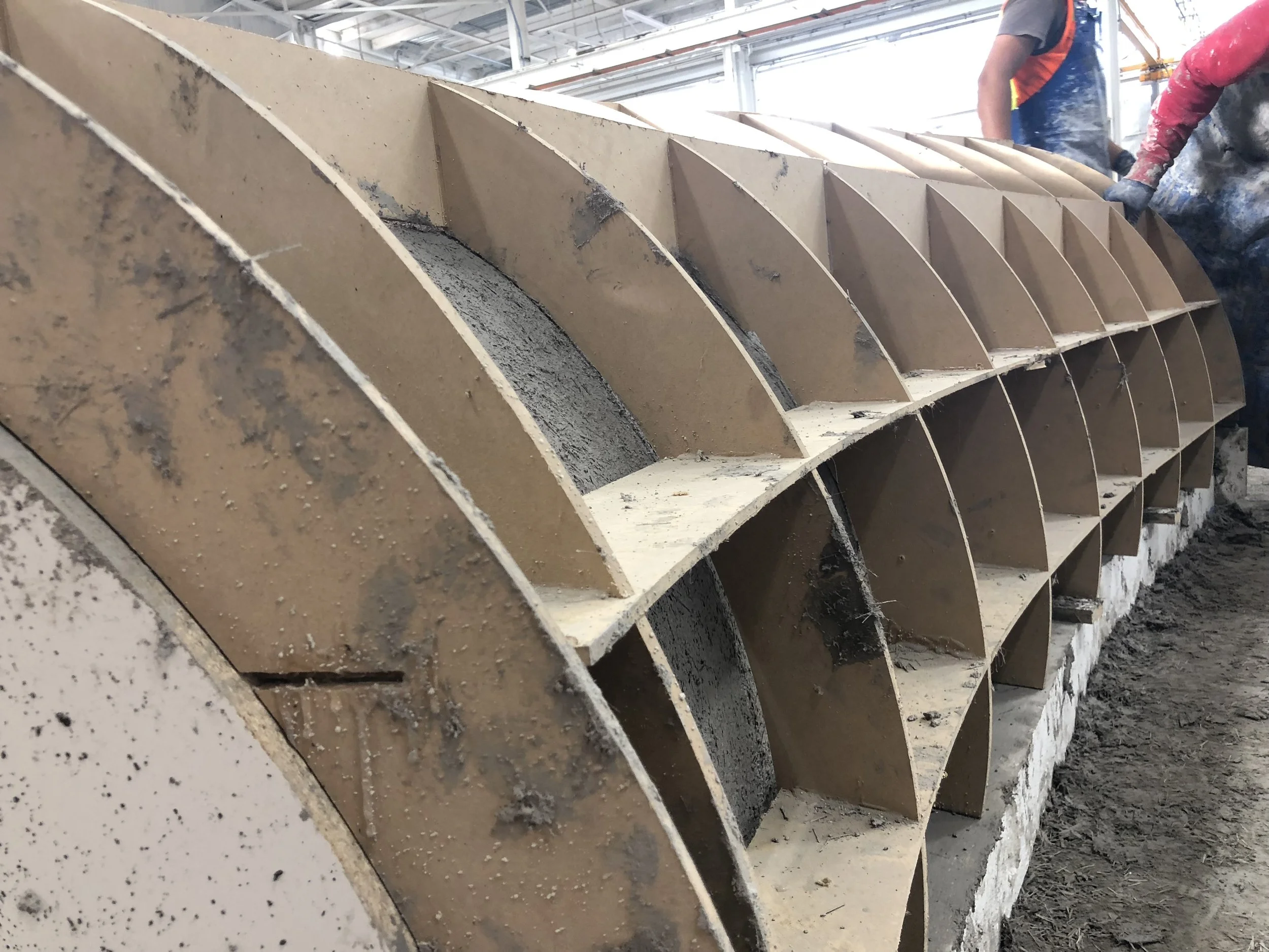Construction workers working on a large, industrial, or construction project inside a building with a metal roof. They are engaged with a big, curved, structural component, possibly made of concrete or similar material.