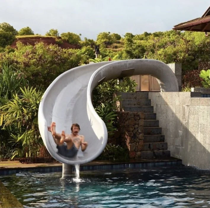 A person sliding down an outdoor water slide into a pool surrounded by lush greenery and stone stairs in the background.