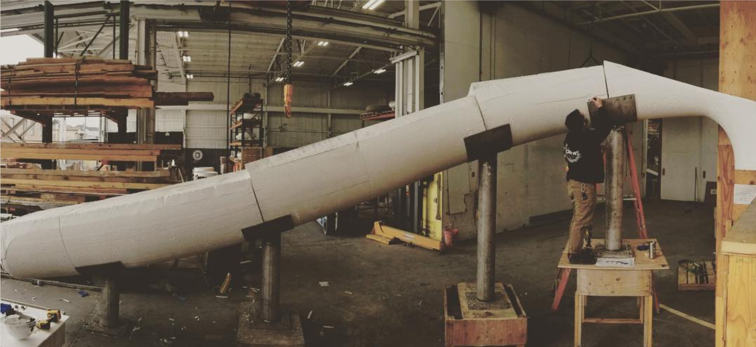 A person in a craft workshop working on the shell of a large rocket or missile, with stacks of wooden planks and equipment around.