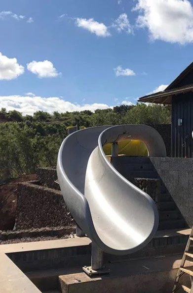 A metal spiral slide at an outdoor play area, with a blue building, green trees, and a partly cloudy sky in the background.