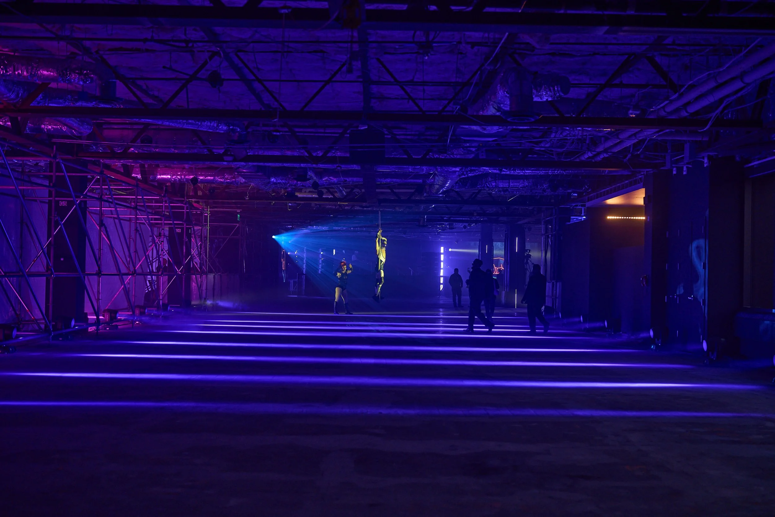 Interior view of a dark space with purple and blue lighting, showing a stage with performers and a large structure made of metal pipes on the left side.
