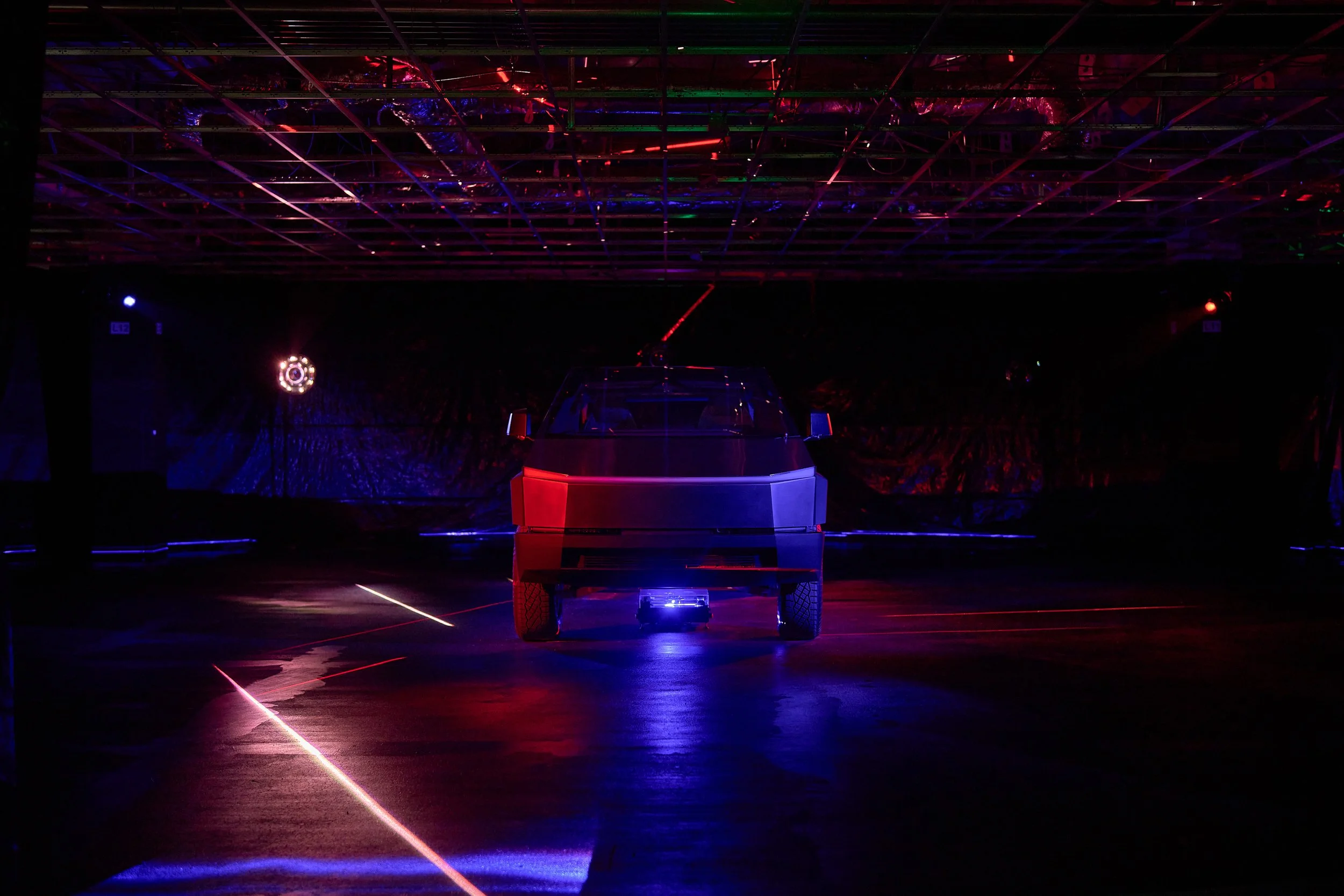 A futuristic, illuminated vehicle is in the center of a dark room with colorful laser lights and reflections on the floor and ceiling.