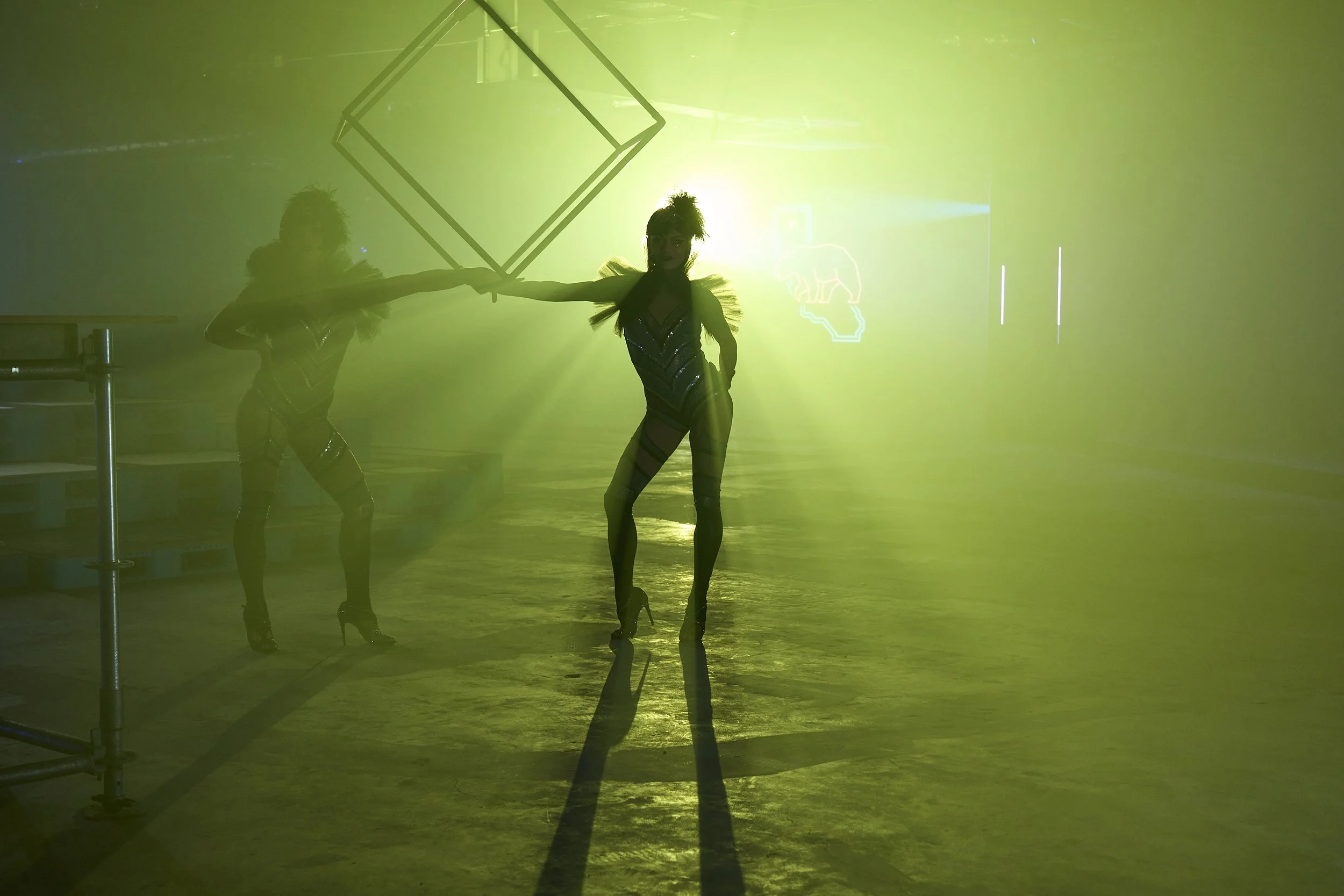 Two performers in futuristic costumes and high heels are on stage in a misty, yellow-green lit environment, holding a large geometric frame between them.