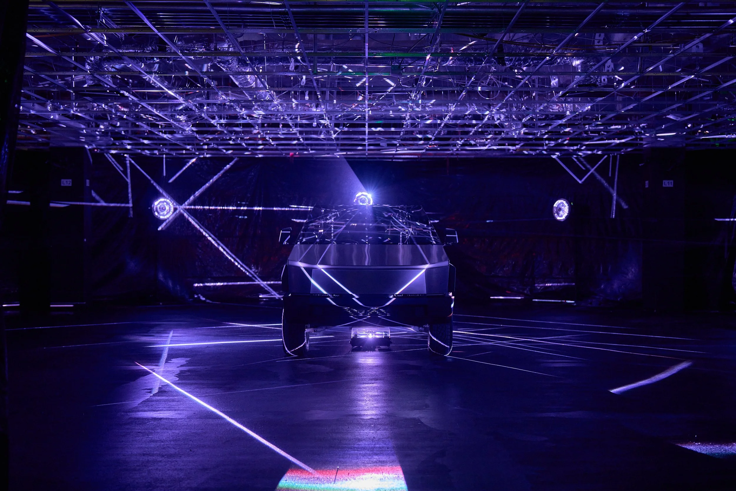 A futuristic, dark room illuminated by vibrant purple and blue lights, with a central structure and laser-like light reflections on the floor.