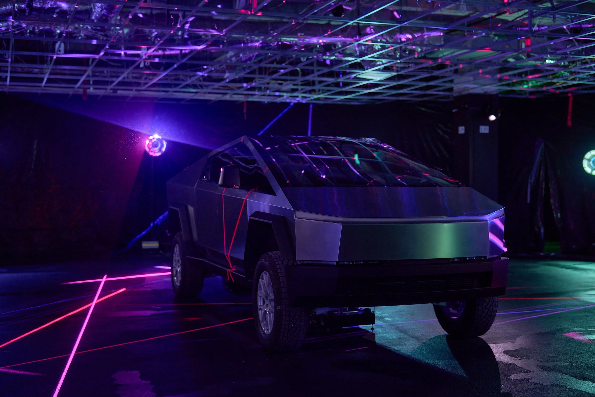 A futuristic black vehicle with sharp angles is illuminated by colorful laser lights in a dark room, creating vibrant reflections on its surface.