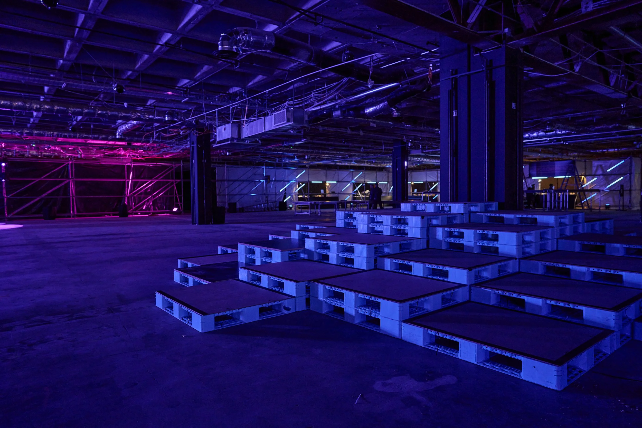 An industrial space decorated with purple and blue neon lights, featuring stacks of pallets and scaffolding, with a person standing in the background.