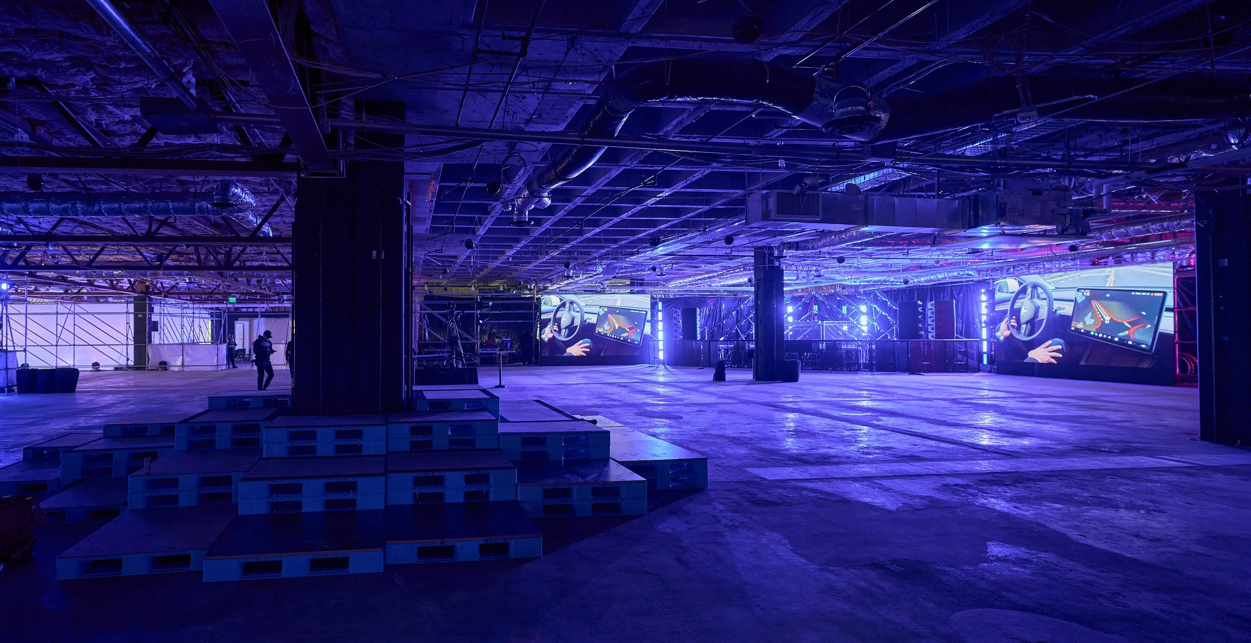 A large indoor space with purple and blue lighting, featuring multiple digital screens showing a car dashboard and steering wheel, with construction materials and scaffolding around.