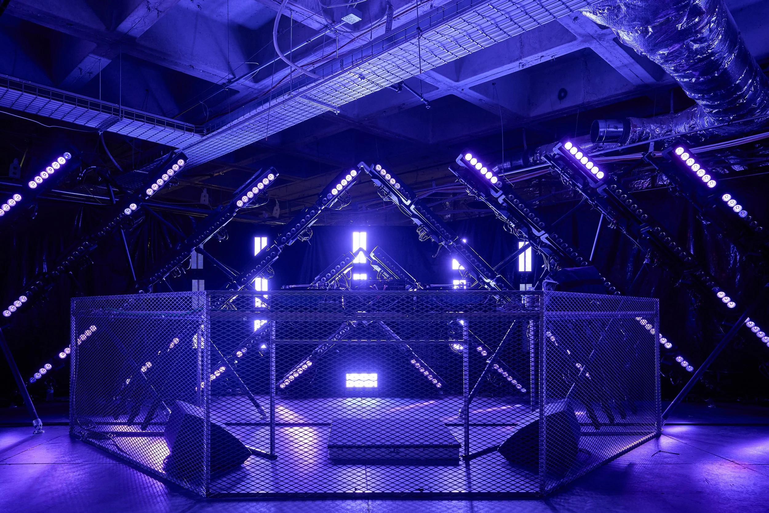 Stage with purple and blue LED lighting, metal scaffolding, safety fencing, and industrial ductwork overhead, likely set up for a concert or performance.