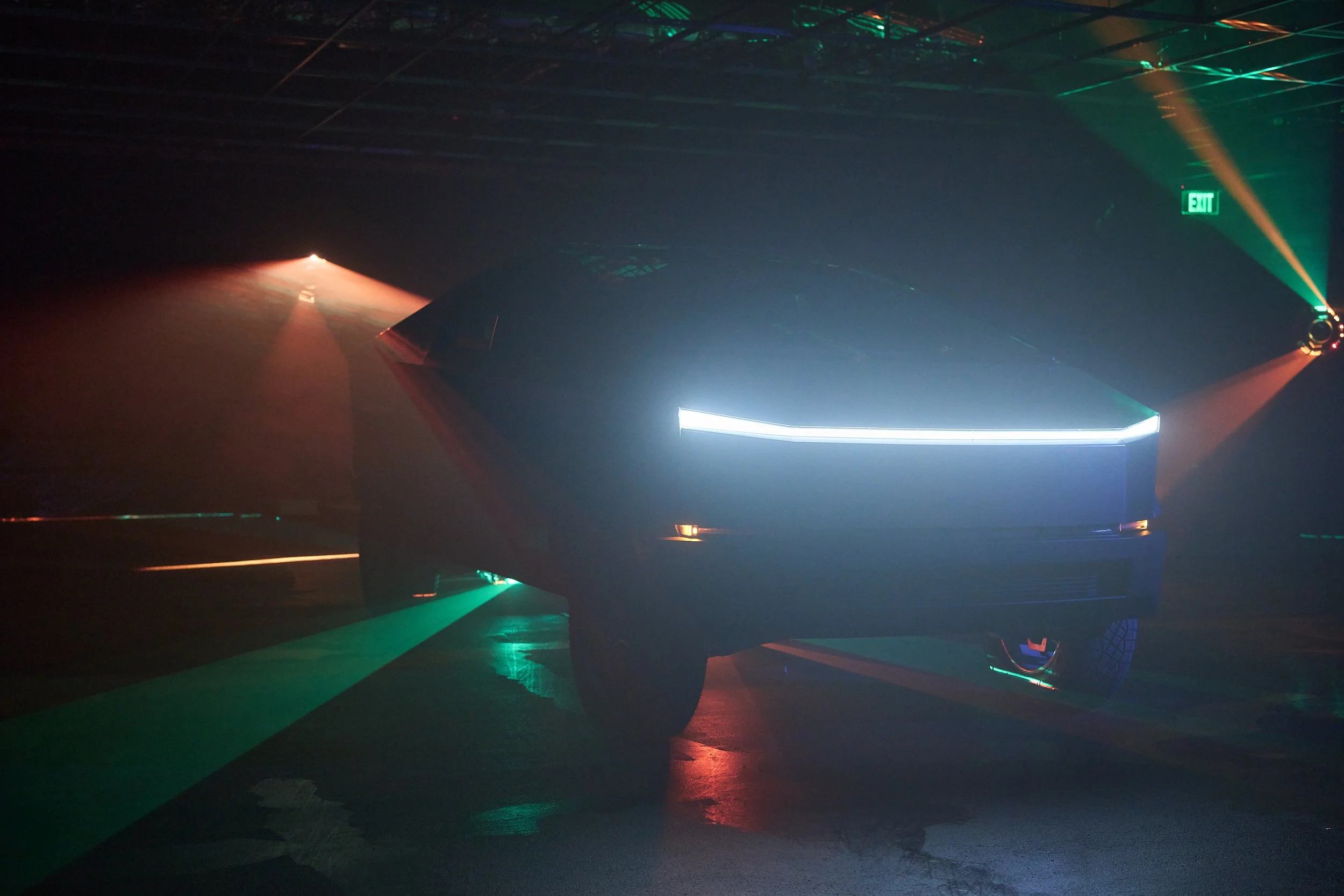 A dimly lit indoor space with laser lights projecting colorful beams on the ceiling and floor, and a dark vehicle, possibly a motorcycle, with illuminated accents visible in the background.