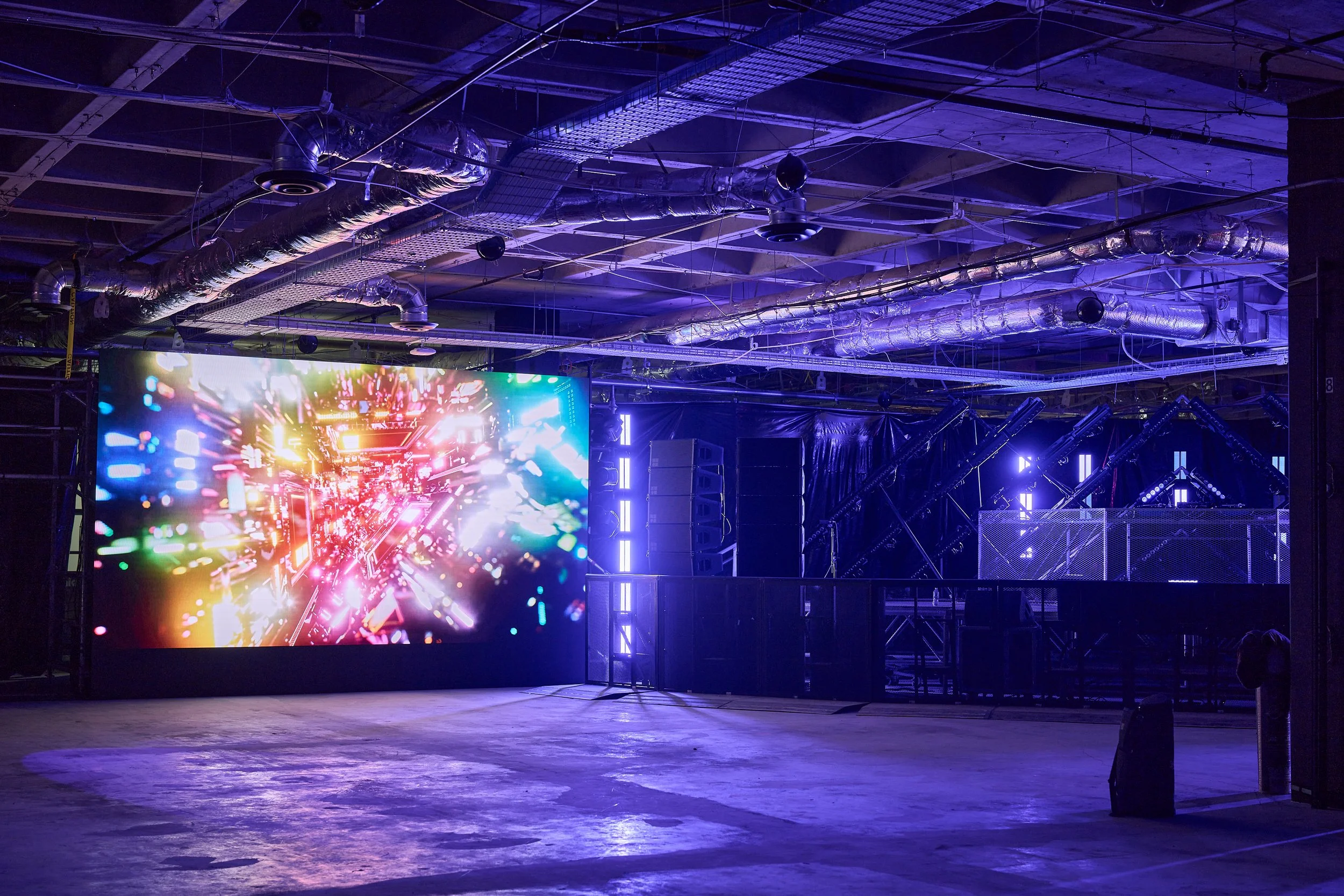 Indoor stage setup with large colorful digital screen displaying abstract lights, with metal scaffolding and lighting equipment overhead, and a reflective floor.
