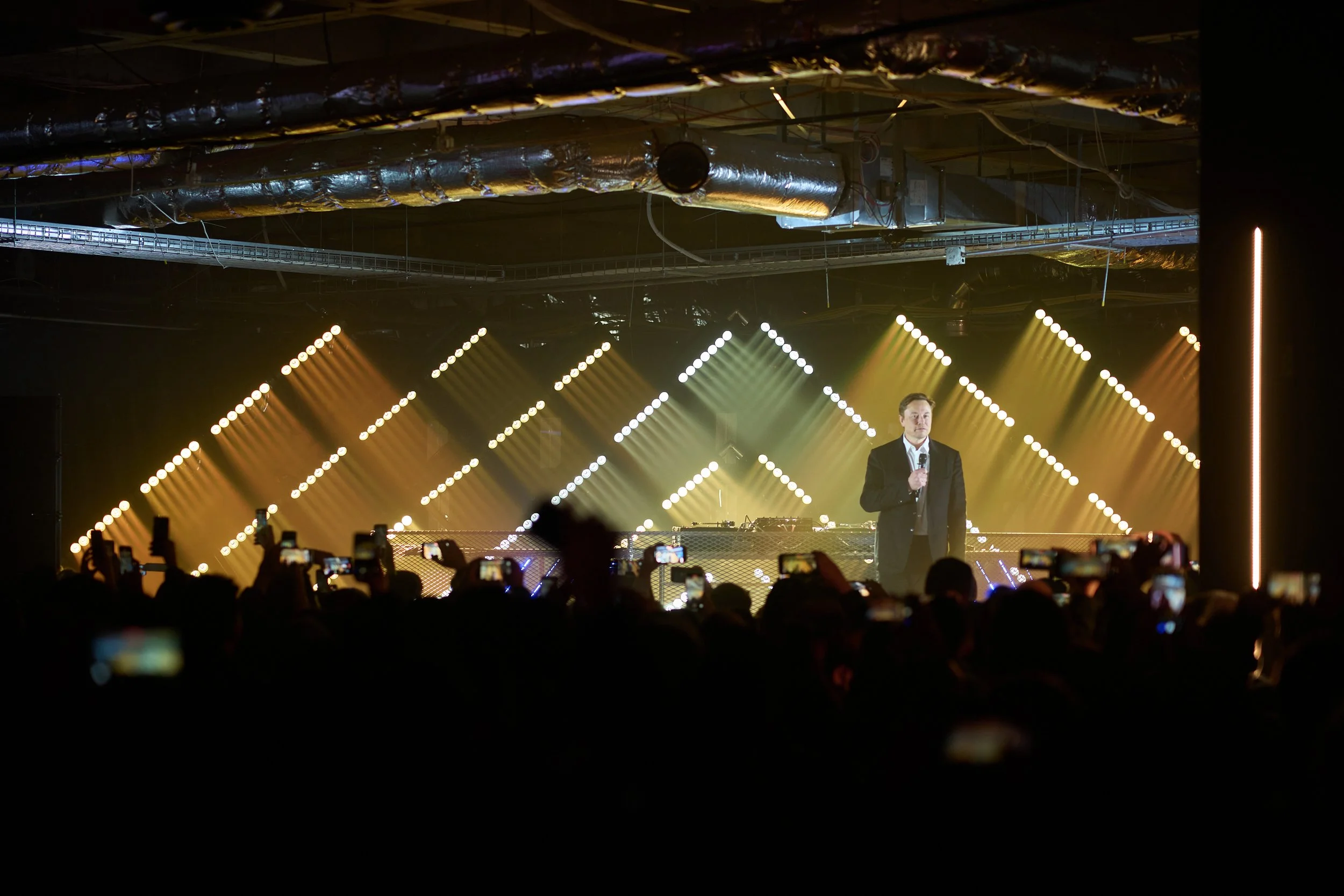 A man in a black suit holding a microphone on stage with colorful stage lights and an audience taking pictures.