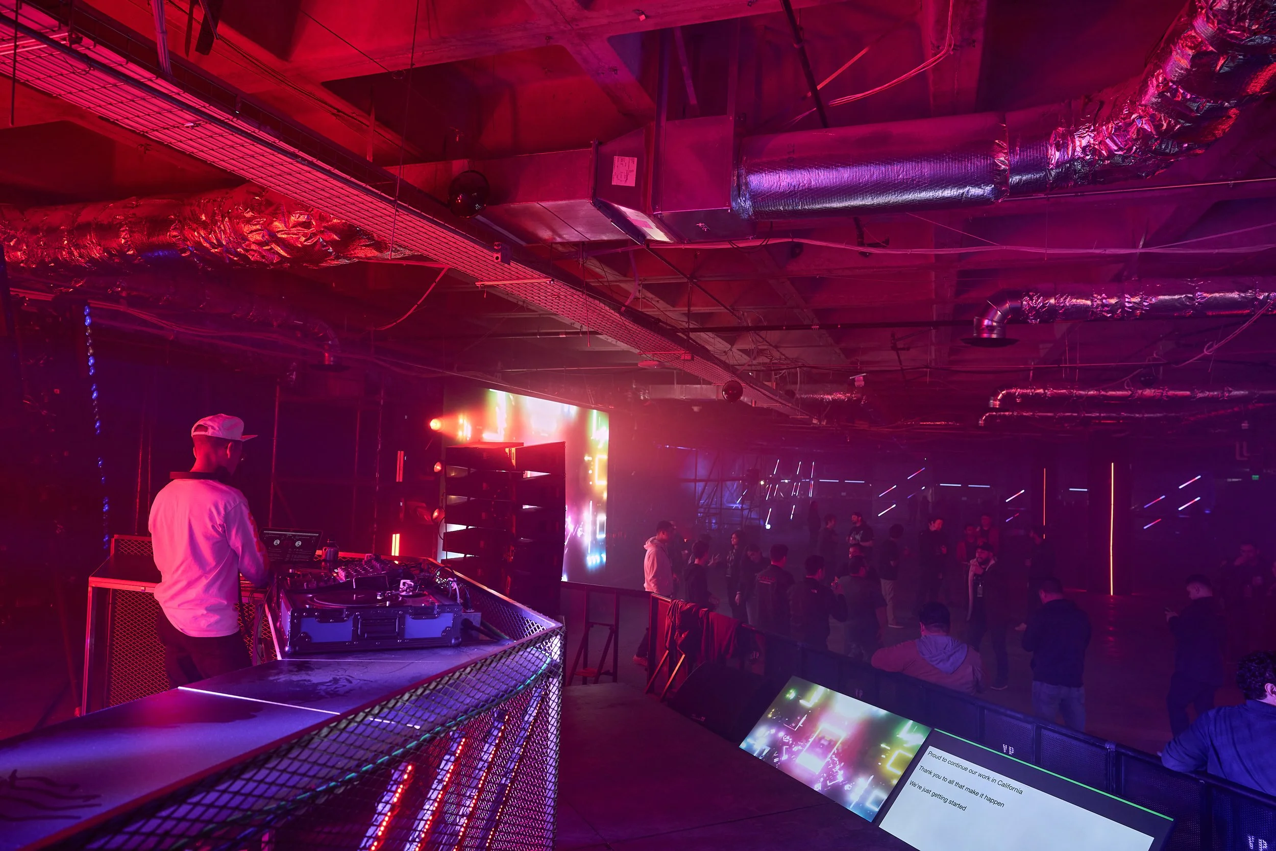 A DJ is performing at a nightclub with dim lighting, colorful neon lights, and a crowd of people dancing underneath an exposed ceiling with visible ducts and pipes.