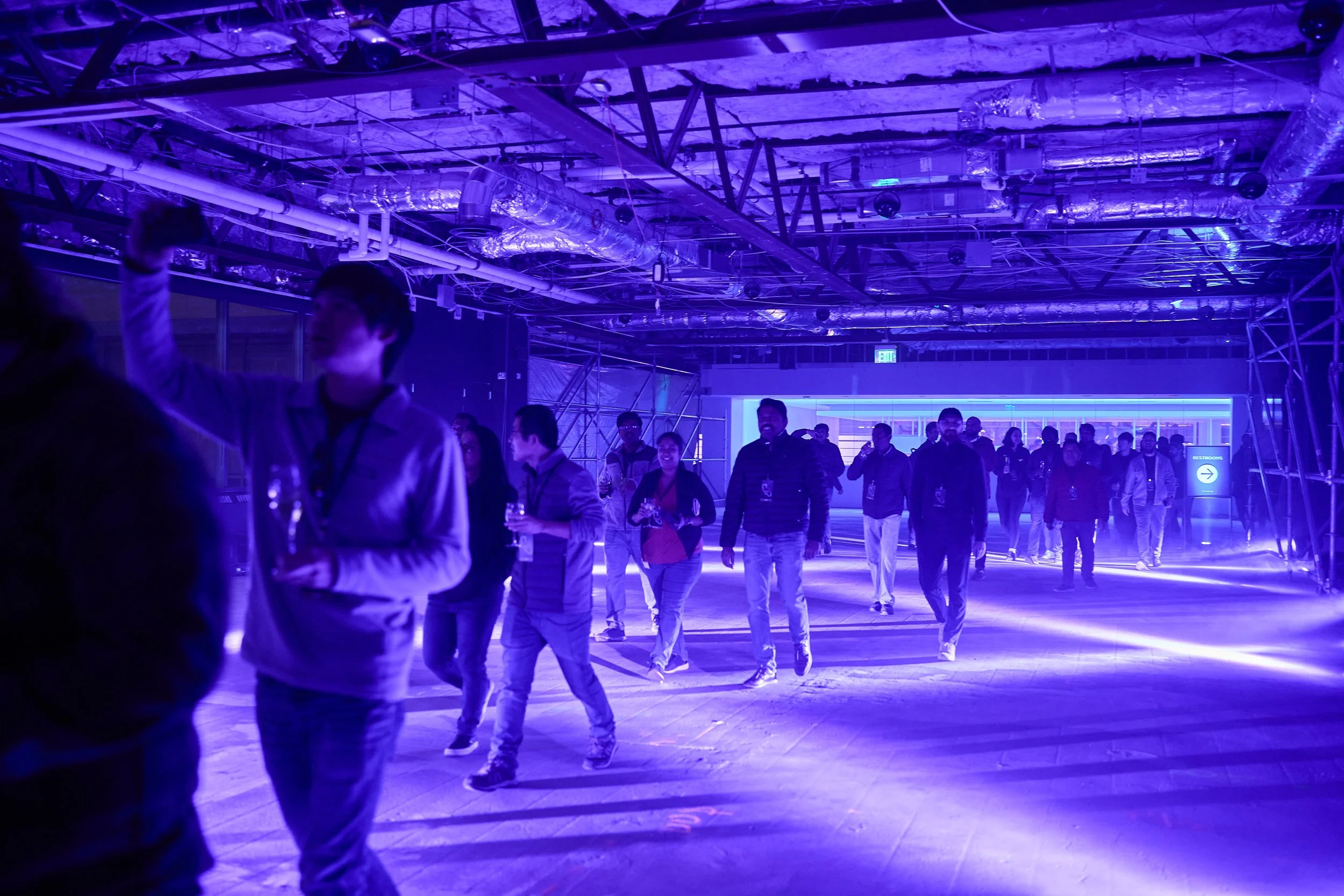 Group of people walking in a dimly lit indoor space with purple lighting, exposed ceiling pipes, and scaffolding structure.