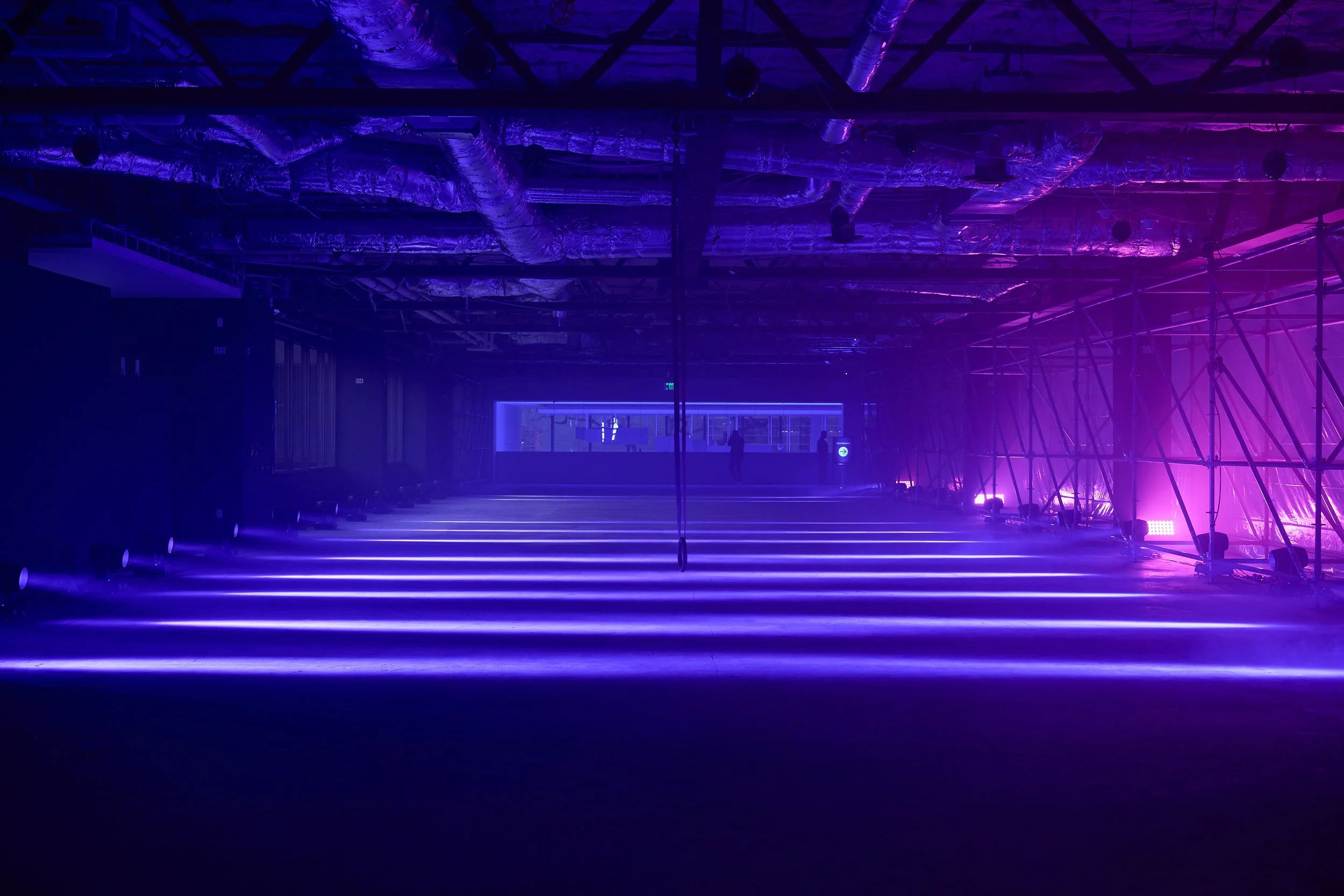 A dark indoor space with purple and blue lighting, featuring stage lights on the floor, scaffolding on the right side, and a person in the distance standing near the wall.