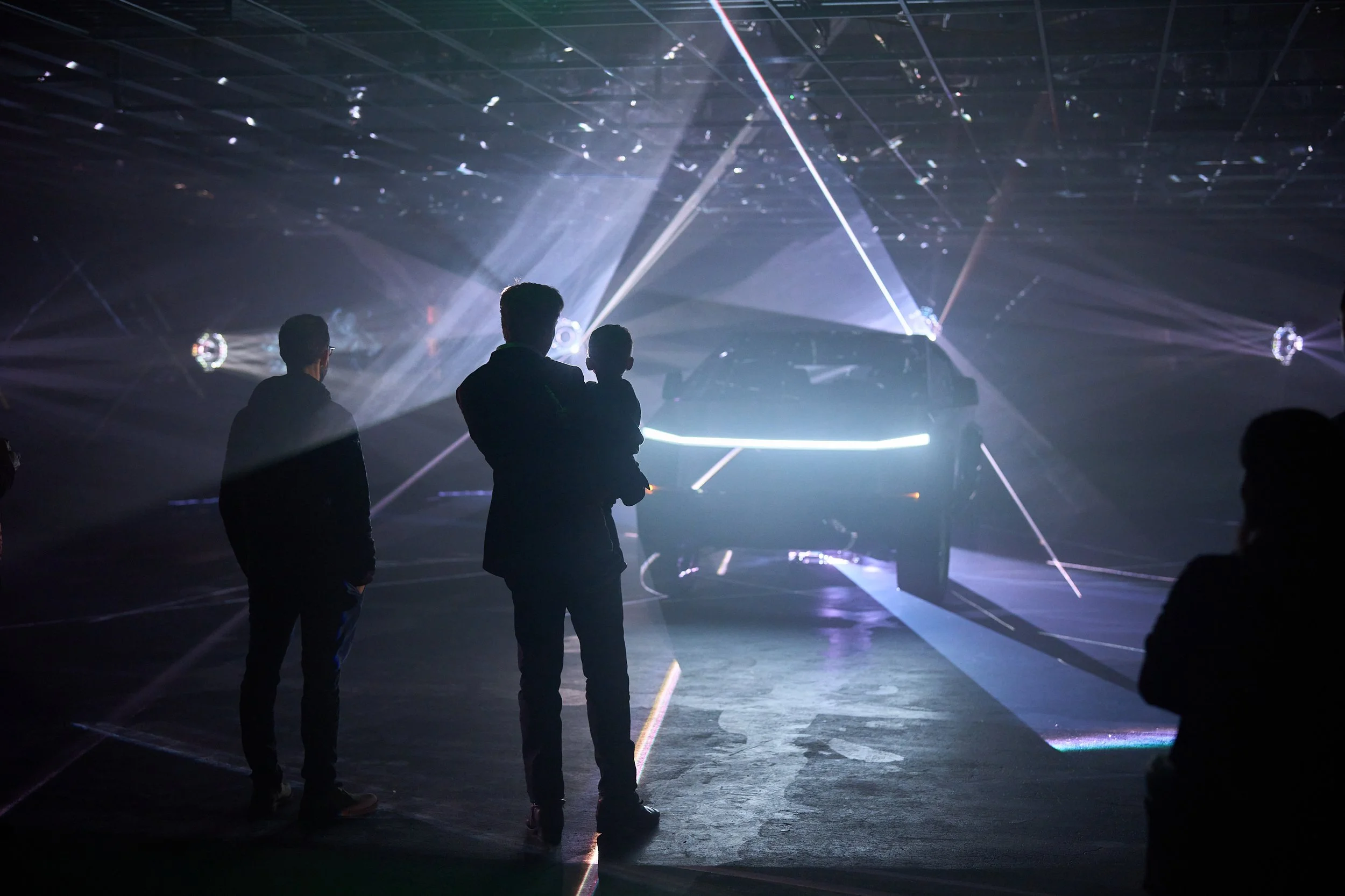 Three silhouettes of people observing a futuristic car surrounded by laser lights and reflections in a dark exhibition space.