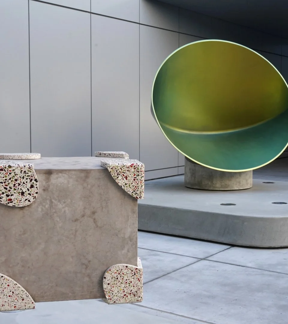 Upside-down terrazzo table with colorful chairs and a green, rounded sculpture on concrete base, modern urban setting