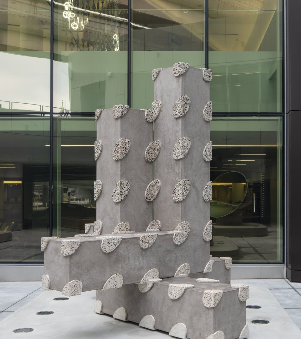 Modern abstract sculpture made of concrete with pebble-like protrusions, situated outside a glass building.
