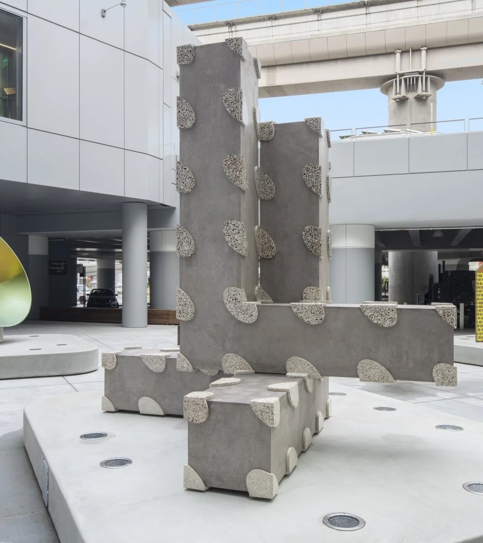Modern outdoor sculpture made of concrete and textured pebble-like elements arranged in vertical and horizontal blocks on a city plaza.