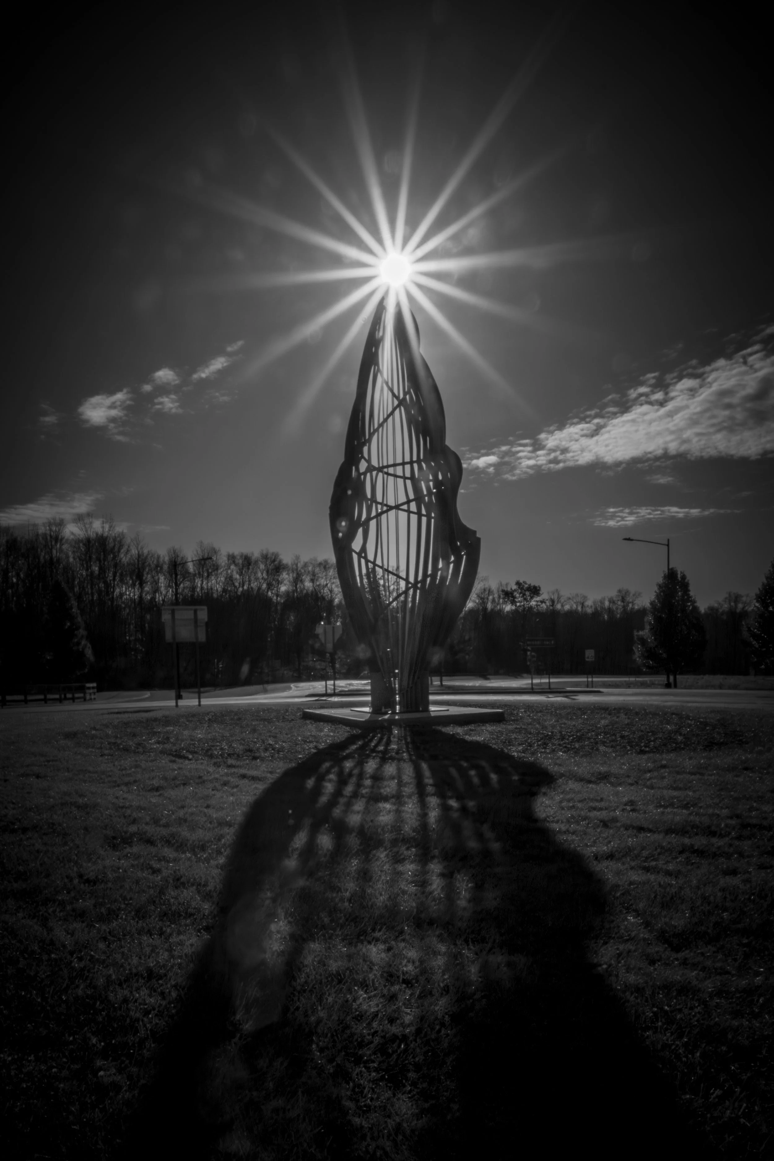 A metal sculpture of a hand reaching upward, with the sun directly behind the fingertips, creating a starburst effect and casting a shadow on the ground. The scene is in black and white, with a partly cloudy sky and trees in the background.