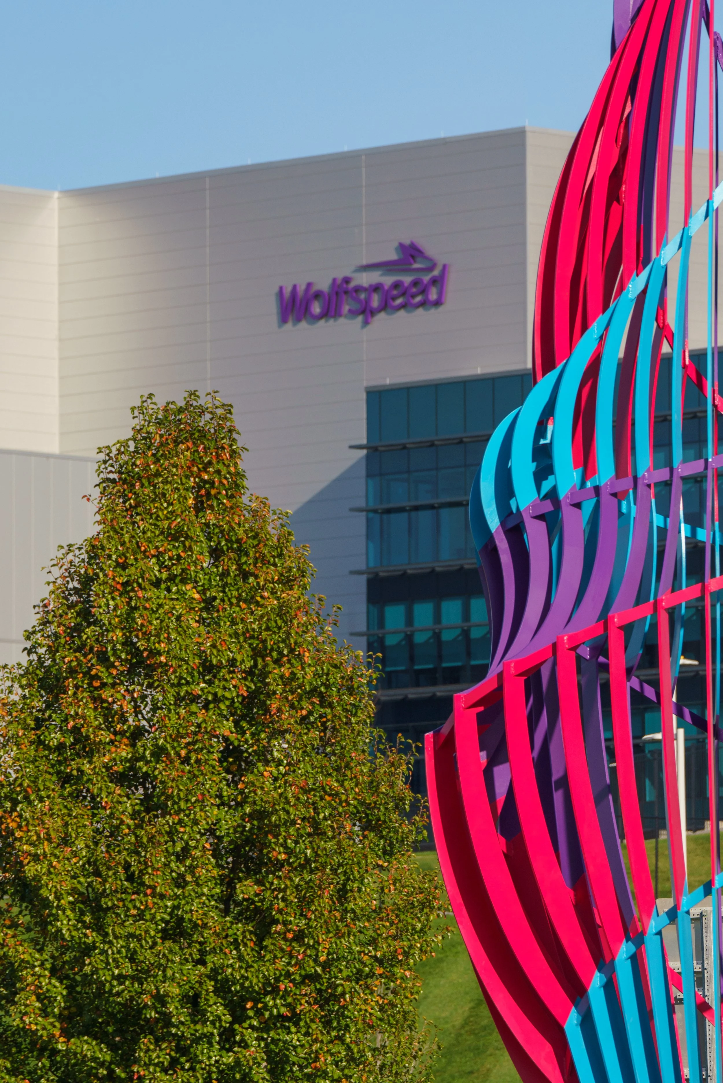 Colorful modern sculpture located outside a building with the 'Wolfspeed' logo against a blue sky, with trees and grass nearby.