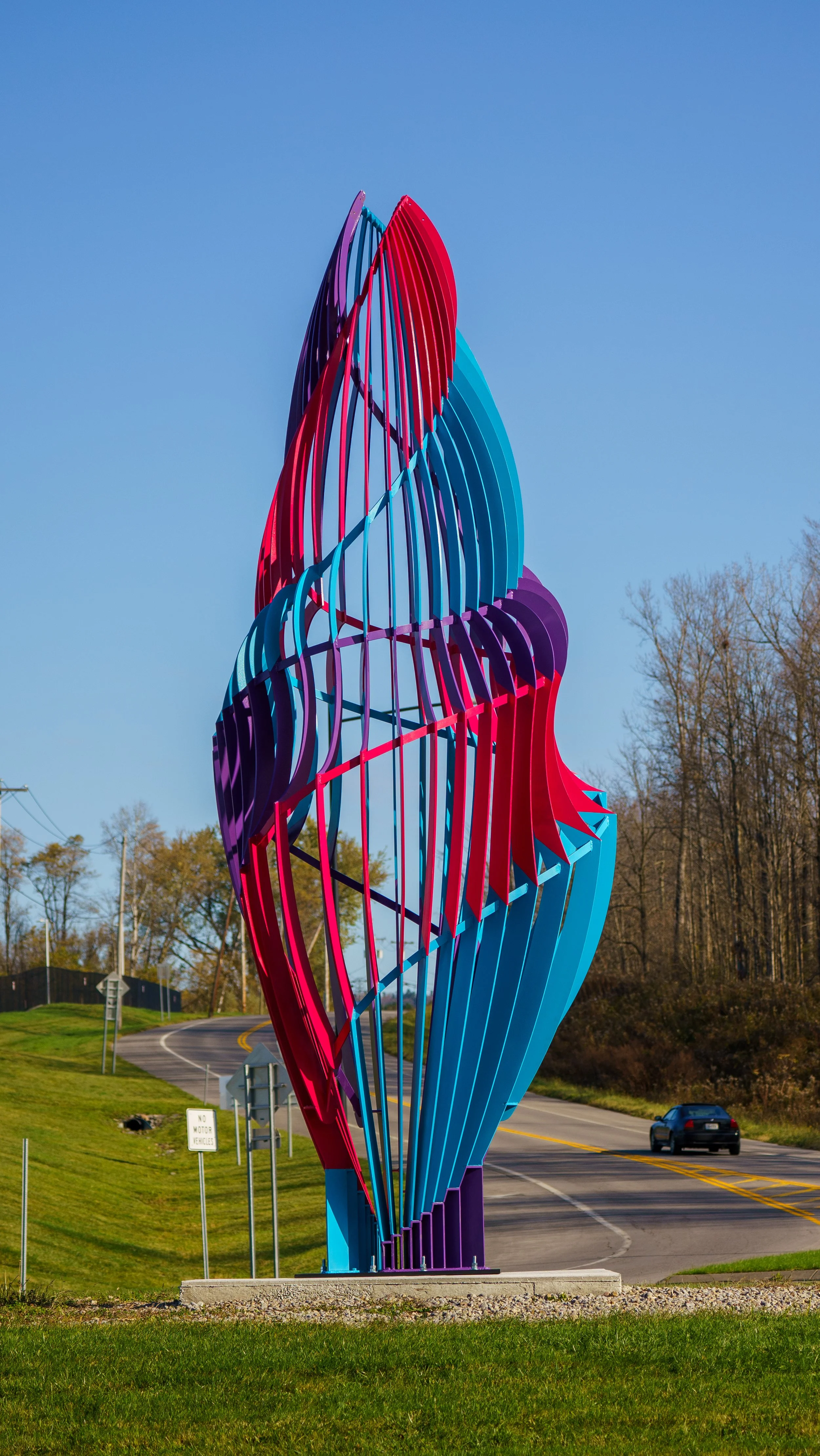 Colorful abstract metal sculpture resembling a flame or flower, with shades of blue, purple, red, and pink, located on a grassy roadside.
