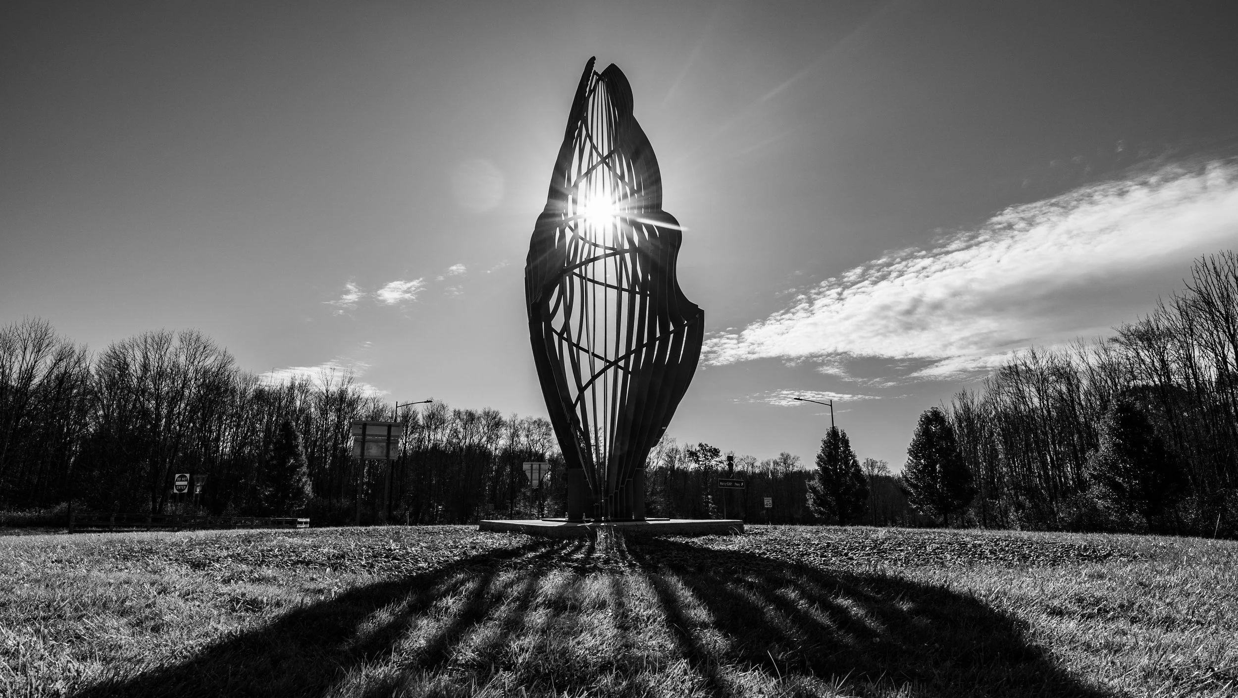 A tall modern sculpture with an open, elongated oval shape, silhouetted against the sun, situated in an outdoor park with trees in the background and shadowed grass in the foreground.