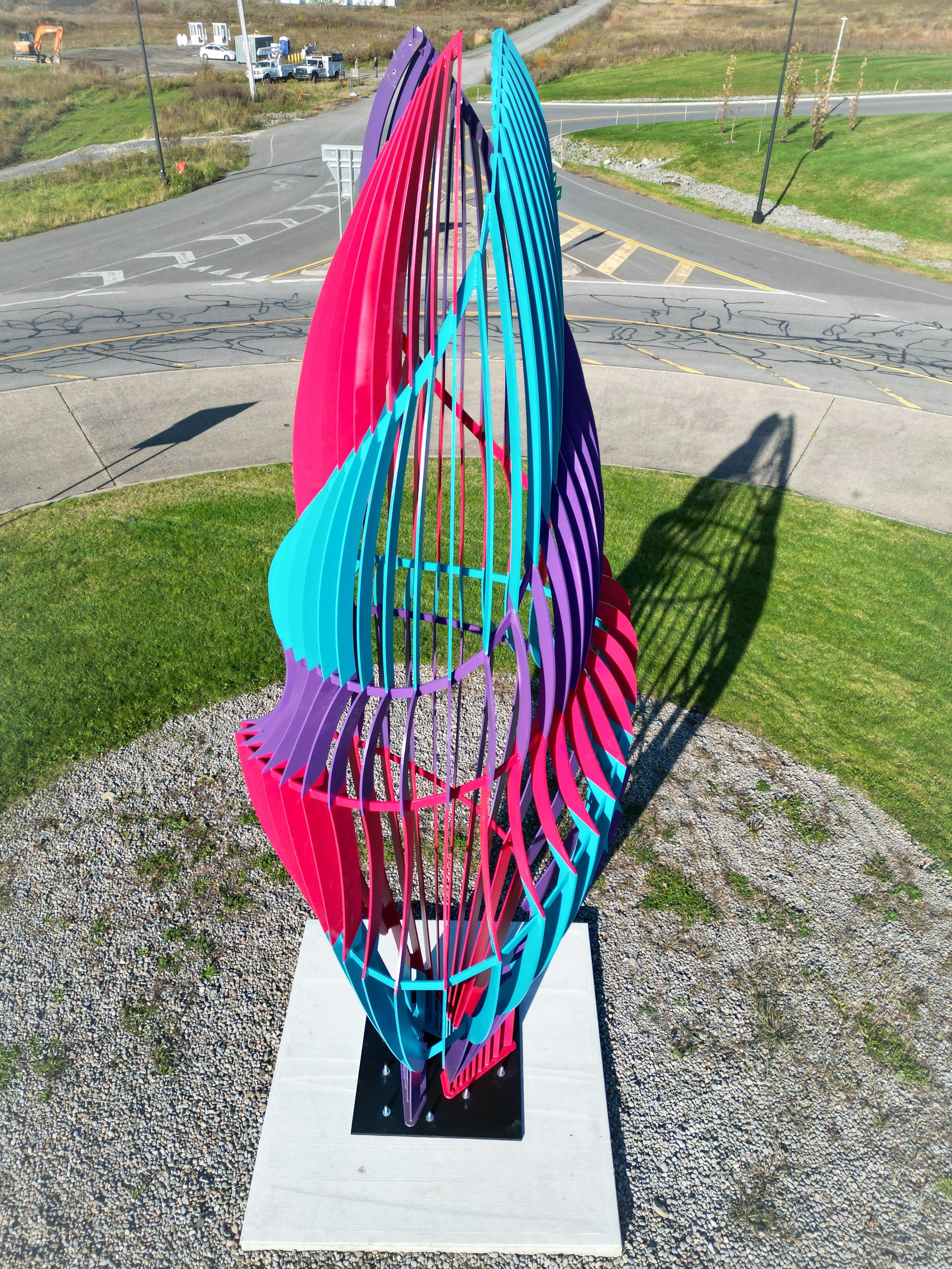 Colorful abstract sculpture consisting of vertically arranged, curved, and interconnected plastic or metal strips in shades of pink, blue, purple, and teal, installed outdoors on a concrete base with gravel around it and shadows cast by the sculpture