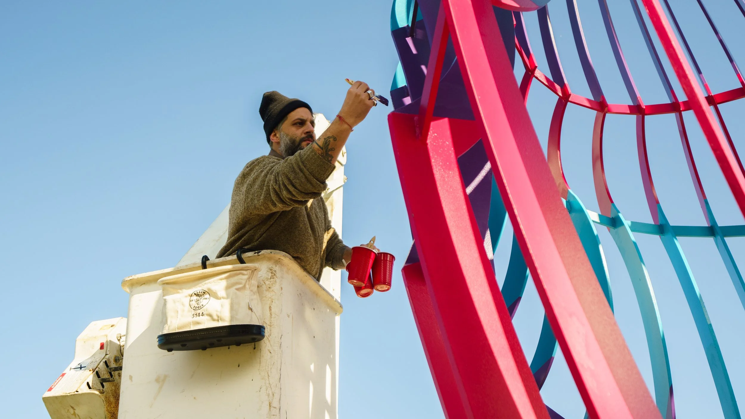 A man painting a colorful Ferris wheel while standing in a white bucket lift against a clear blue sky.