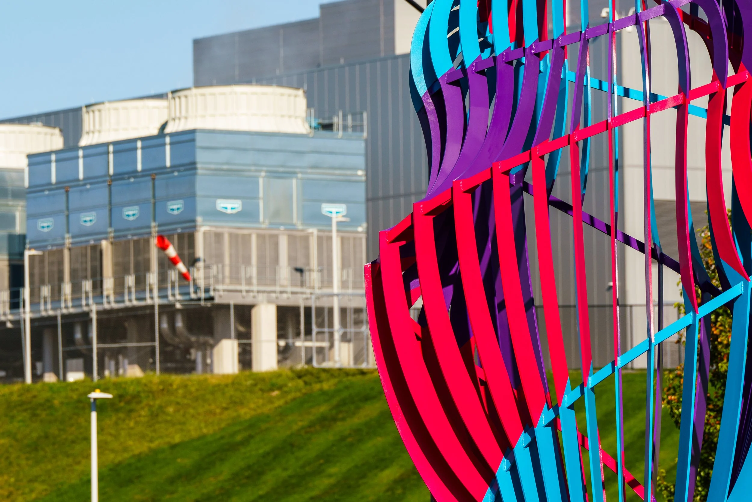 Colorful abstract metal sculpture with purple, red, and blue curved rods, outdoor urban background with modern building, green grass, and blue sky.