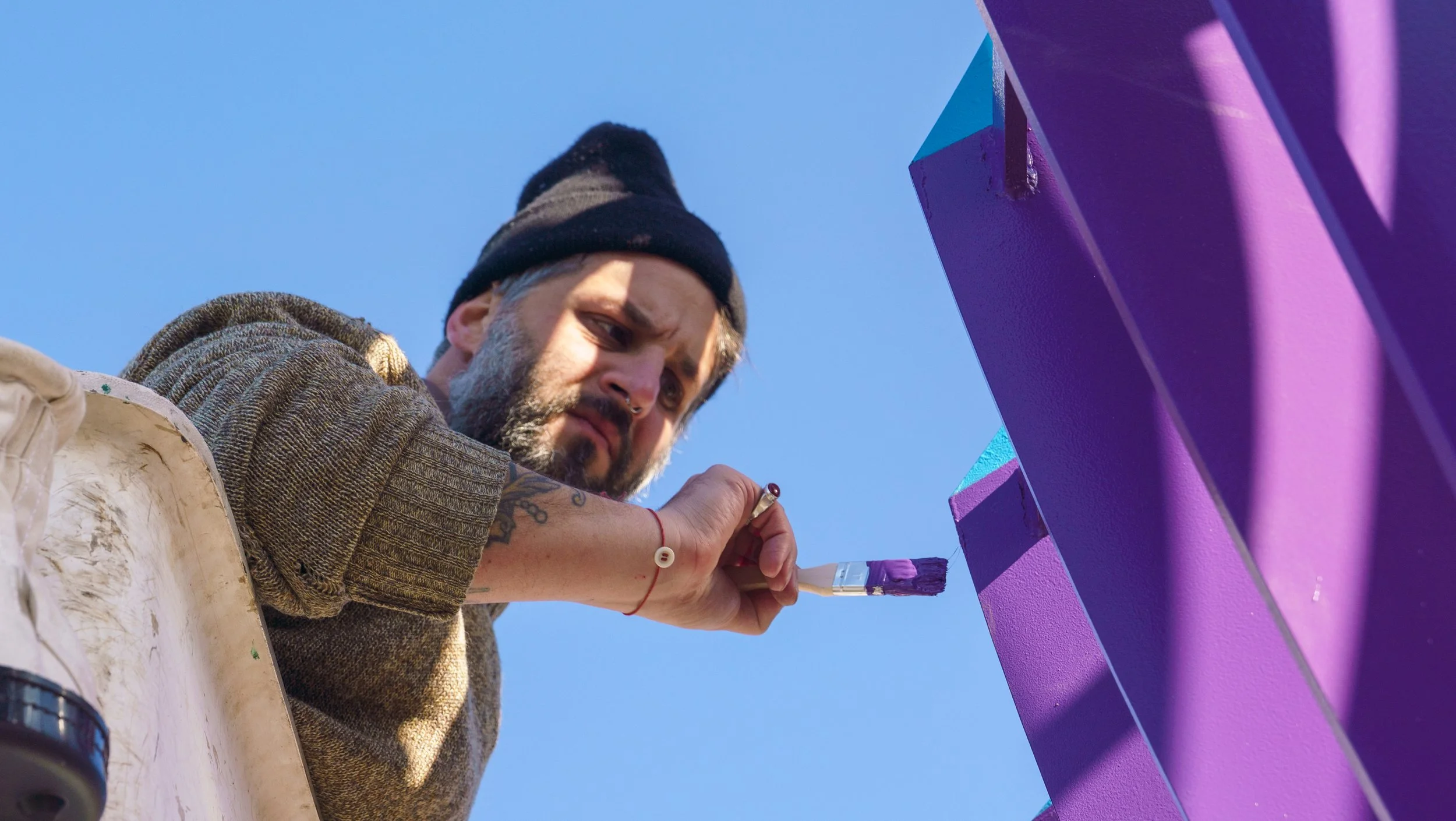 A man wearing a beanie and a brown jacket painting a structure purple with a paintbrush. The sky is clear and blue.