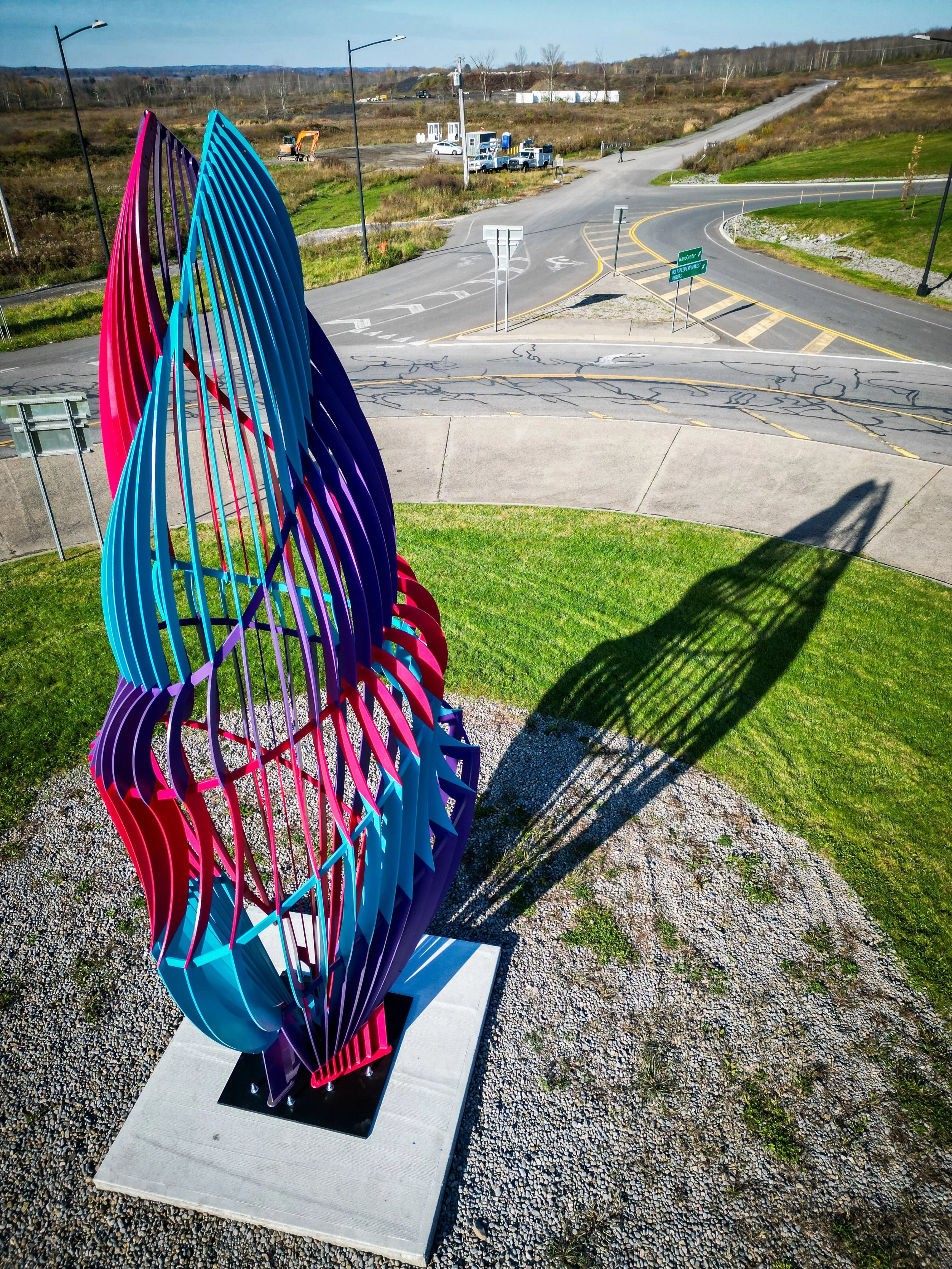 Colorful abstract sculpture on a concrete base with a shadow cast on the ground, set against a rural roadside landscape with a curved road, grassy areas, and trees in the distance.