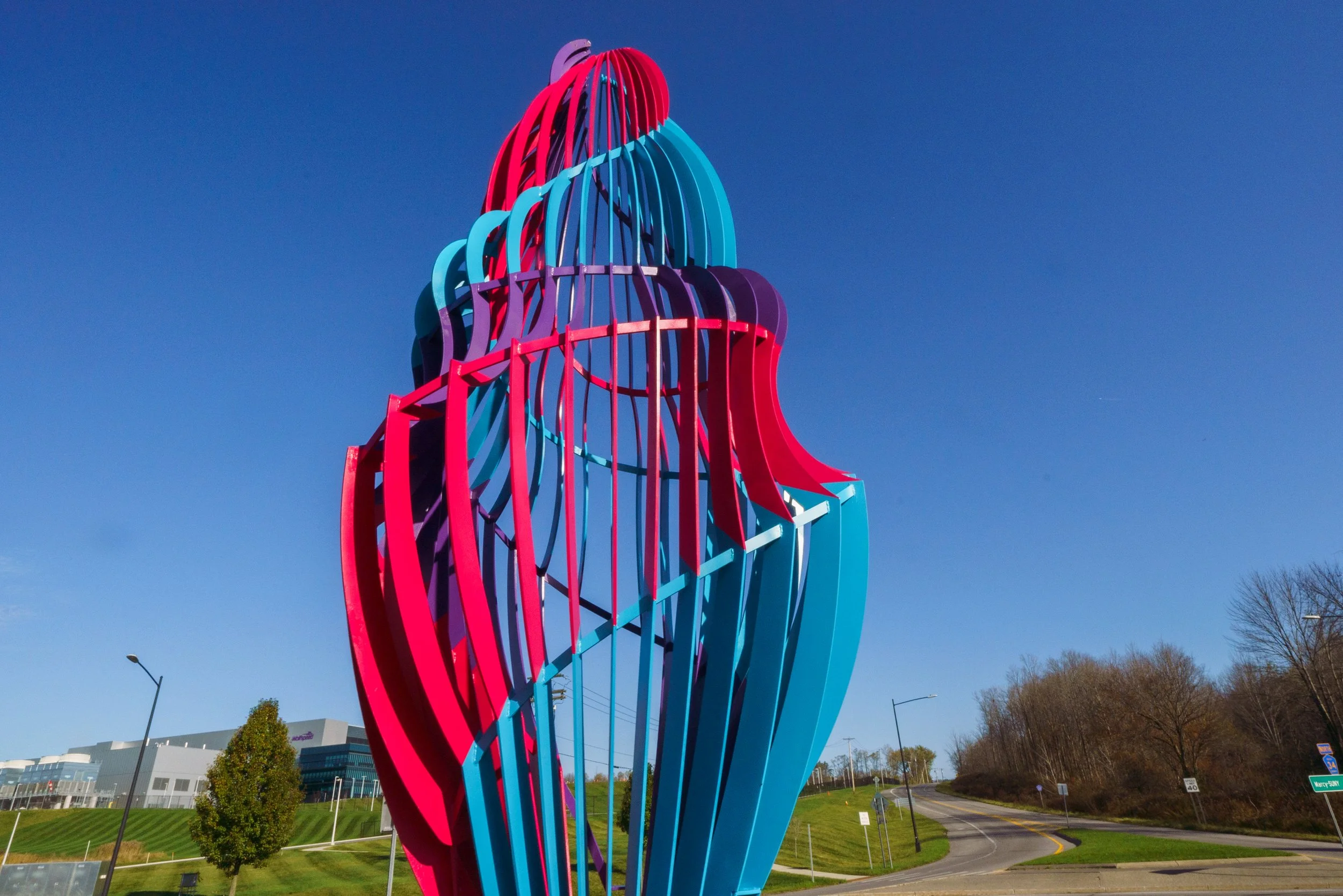 Colorful abstract outdoor sculpture with red, purple, and teal curved ribbons against a blue sky, located near a grassy area, road, and commercial buildings.