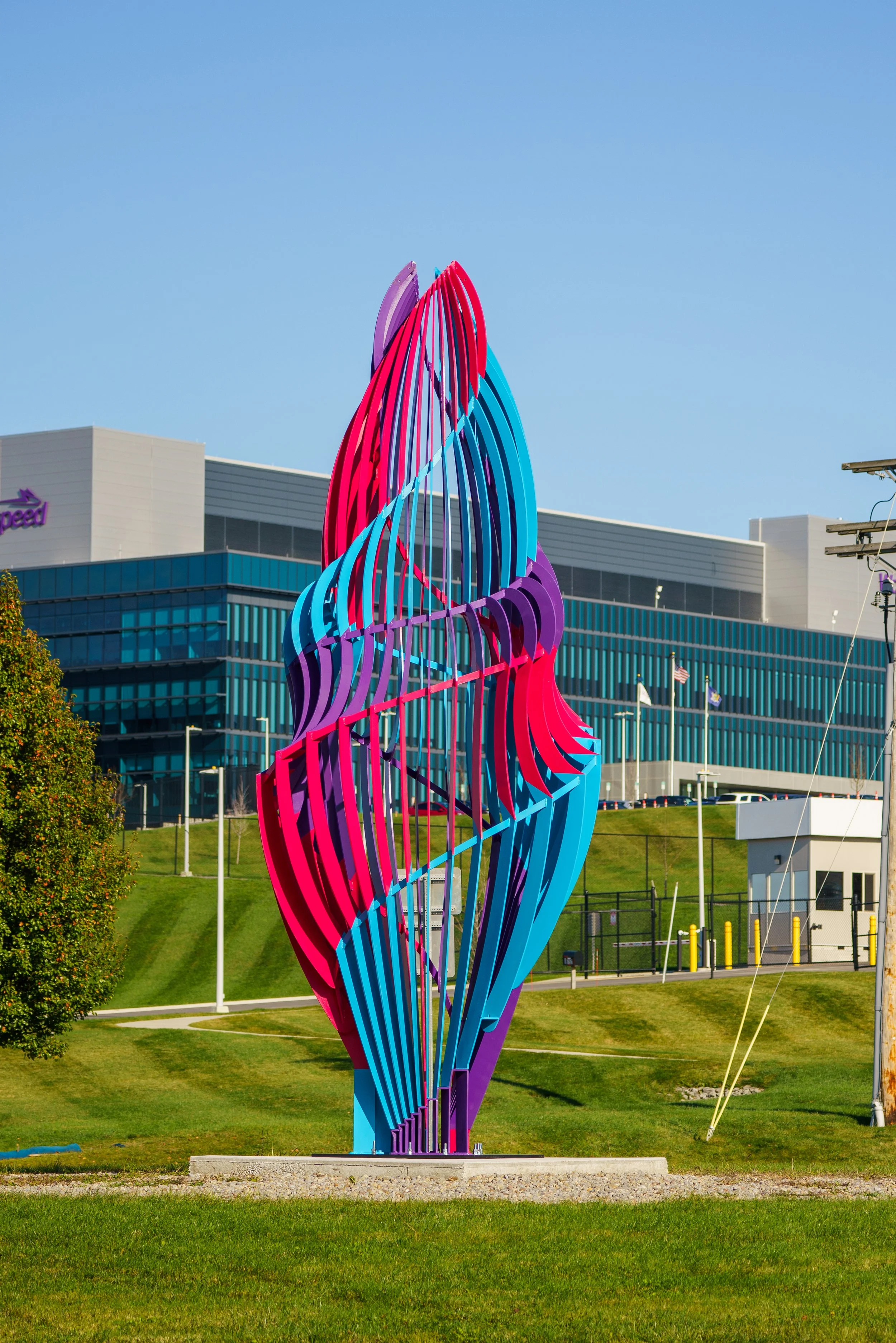 Colorful abstract outdoor sculpture with curved, elongated shapes in pink, purple, blue, and teal, set on a concrete base in a park with green grass, trees, and modern building in the background under a clear blue sky.