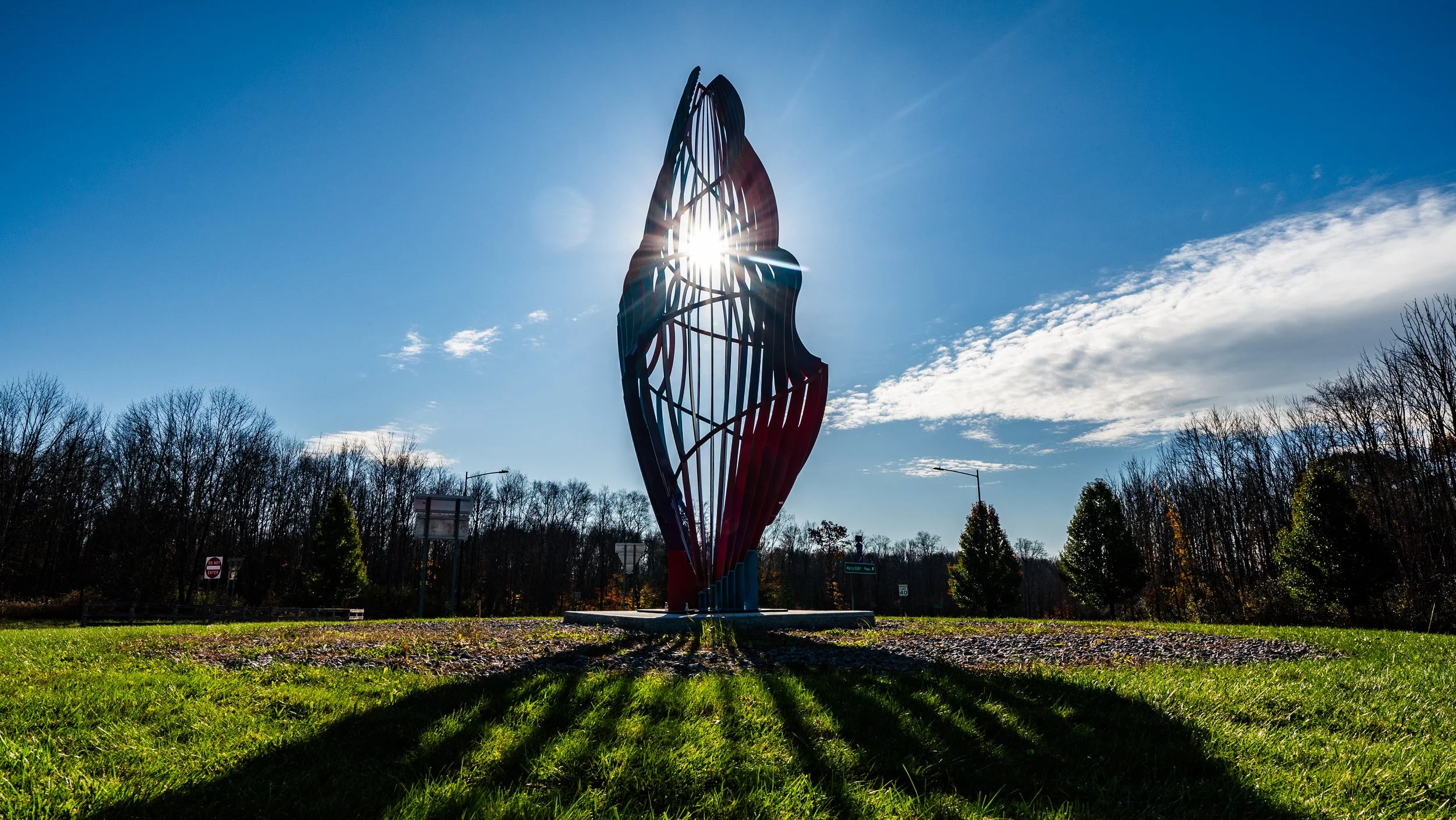 A modern abstract sculpture with a sun shining behind it, creating a glow. The sculpture is positioned in a park with trees and a clear blue sky with some clouds.